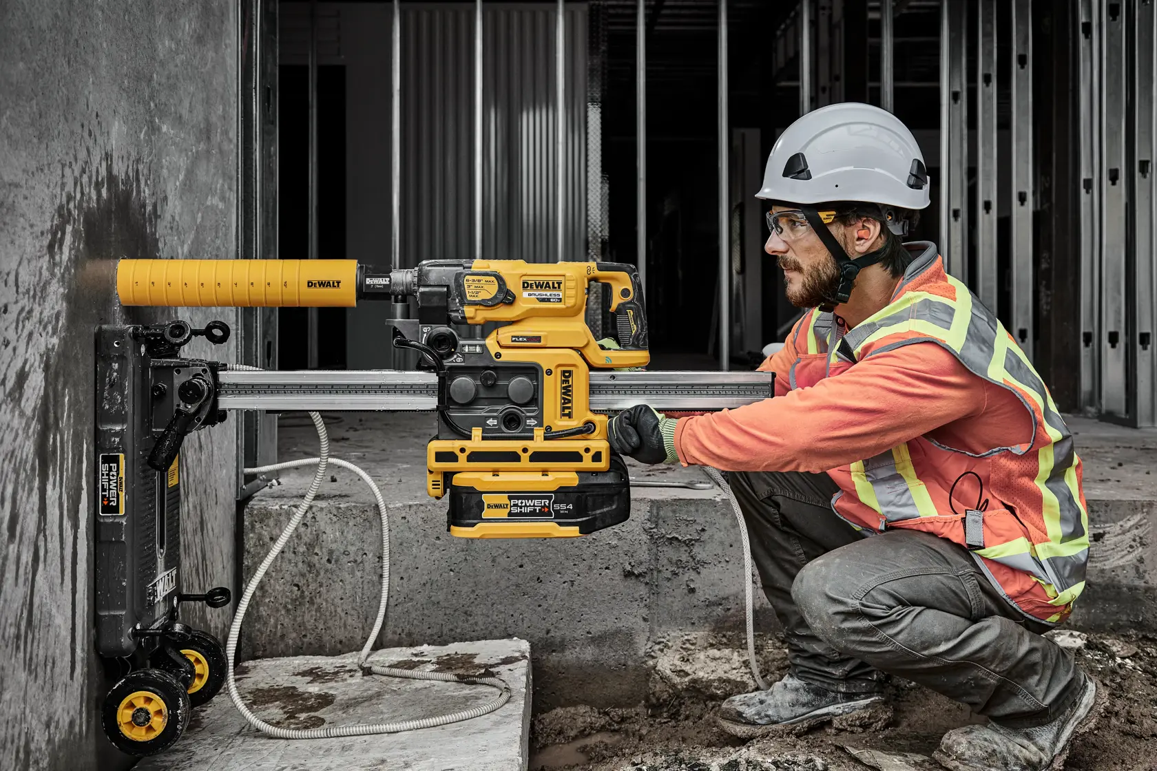 A construction worker wearing a safety helmet and reflective vest uses a yellow DEWALT rotary hammer drill with dust extraction on a concrete wall at an industrial building site.