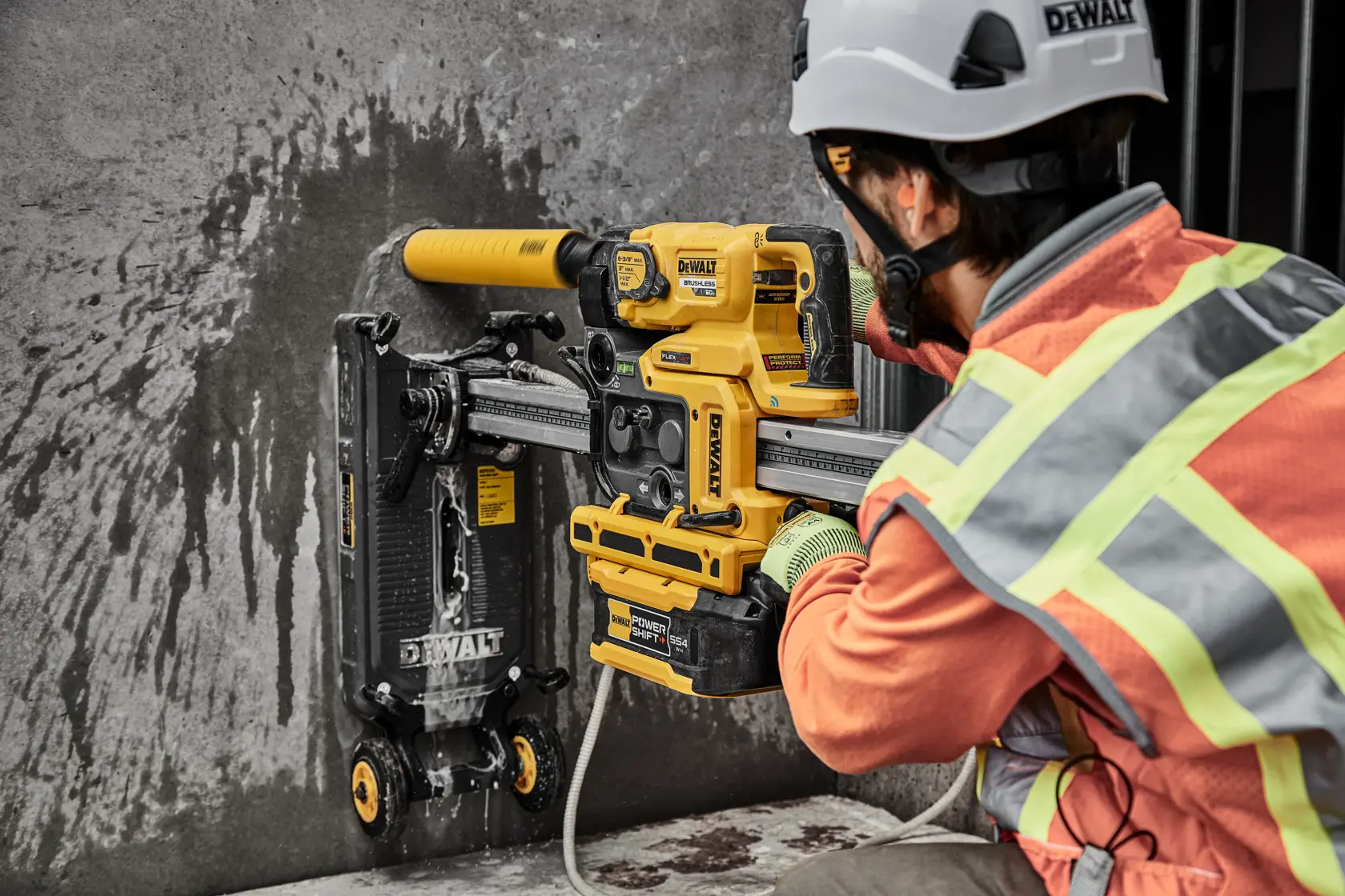 A construction worker wearing a helmet and safety vest operates a yellow DEWALT power tool to drill into a concrete wall. The worker is using a mounted drilling apparatus, and water is visible on the wall surface.
