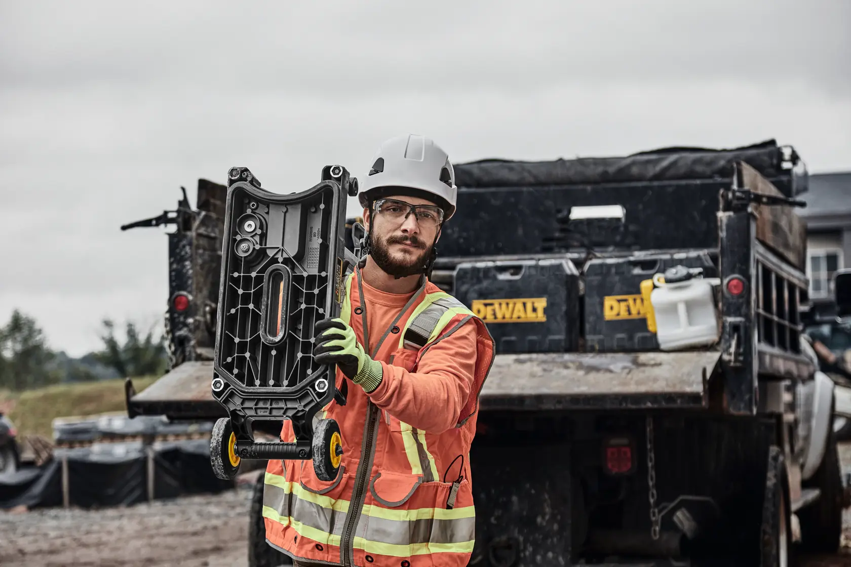 A construction worker wearing a safety vest, gloves, and a helmet is holding a DEWALT tool with wheels on a construction site. The worker is standing in front of a truck with DEWALT branding and other equipment visible in the background.