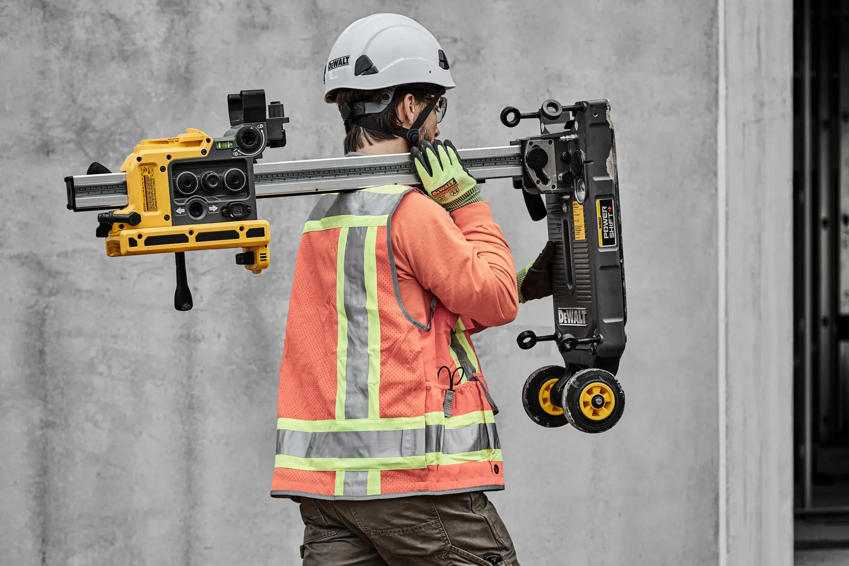 A construction worker wearing a safety helmet, gloves, and a reflective orange vest carries a DEWALT tool with wheels and a yellow component over their shoulder, walking near a concrete wall.