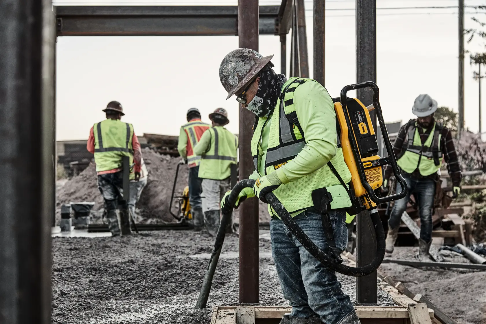 Construction workers wearing safety gear, including helmets and high-visibility vests, are operating DEWALT backpack equipment while working on a concrete site. One worker is using a hose connected to the device, while others are visible in the background.