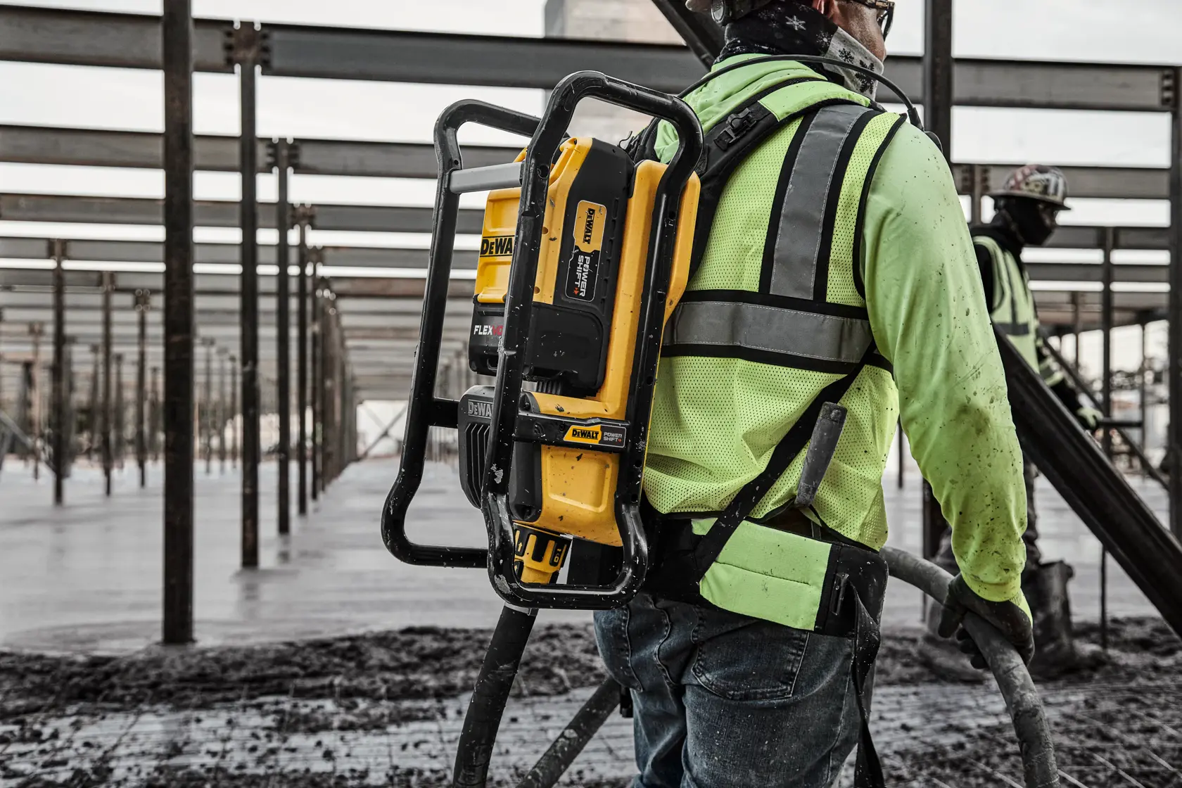 A construction worker wearing a high-visibility safety vest and harness is operating equipment at a construction site. The worker carries a DEWALT device, attached to a metal frame backpack, while handling a hose over freshly poured concrete. The background shows steel beams and another worker in safety gear.