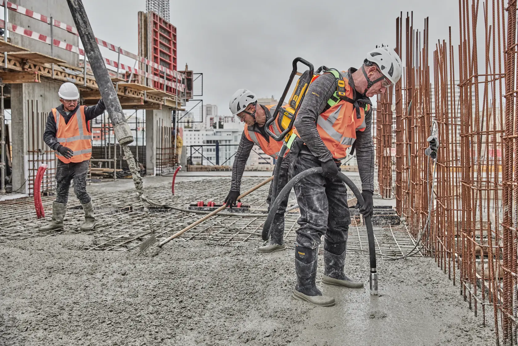 Construction workers wearing safety helmets and orange vests operate a concrete vibrator and pump on a building site, pouring and leveling fresh concrete. Metal reinforcement bars and building materials are visible around them.