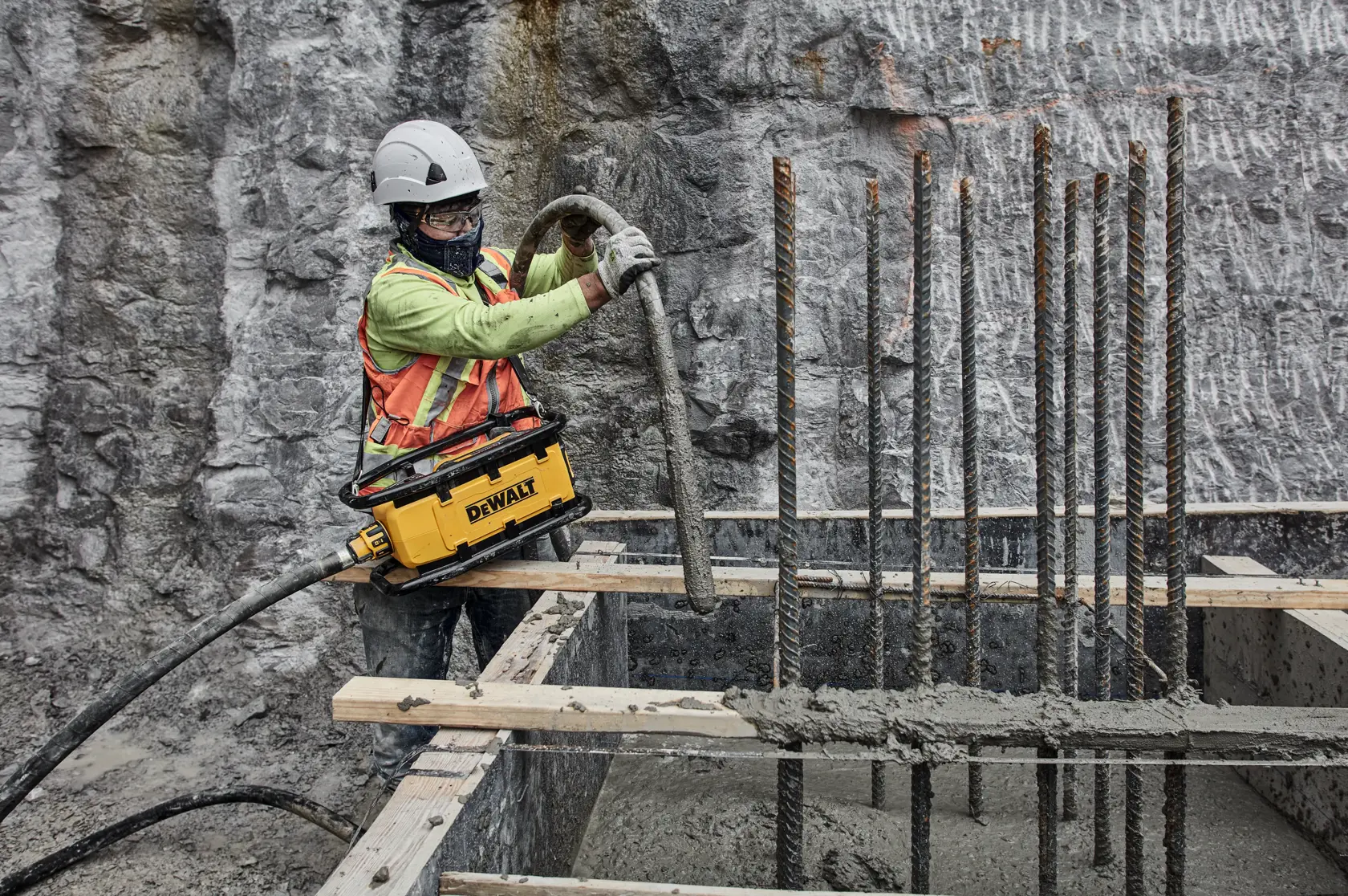 A construction worker wearing a helmet and safety vest operates a DEWALT concrete vibrator tool, pouring concrete into a foundation reinforced with rebar rods at a construction site with a rocky wall in the background.