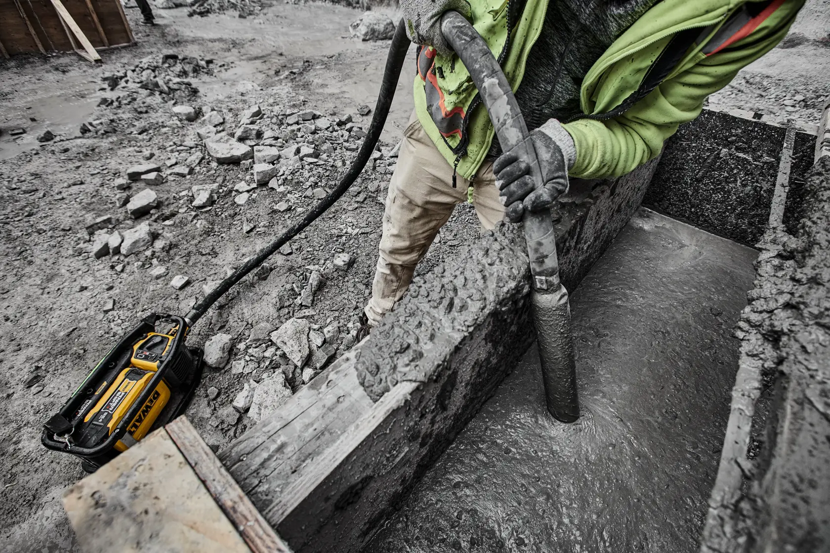 A worker using a DEWALT cordless concrete vibrator on a construction site to smooth out freshly poured concrete in a wooden form.