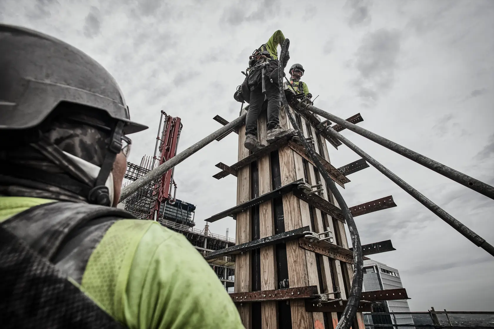 Construction workers wearing safety gear are working at height on a wooden structure with metal supports, surrounded by building equipment and a cloudy sky.