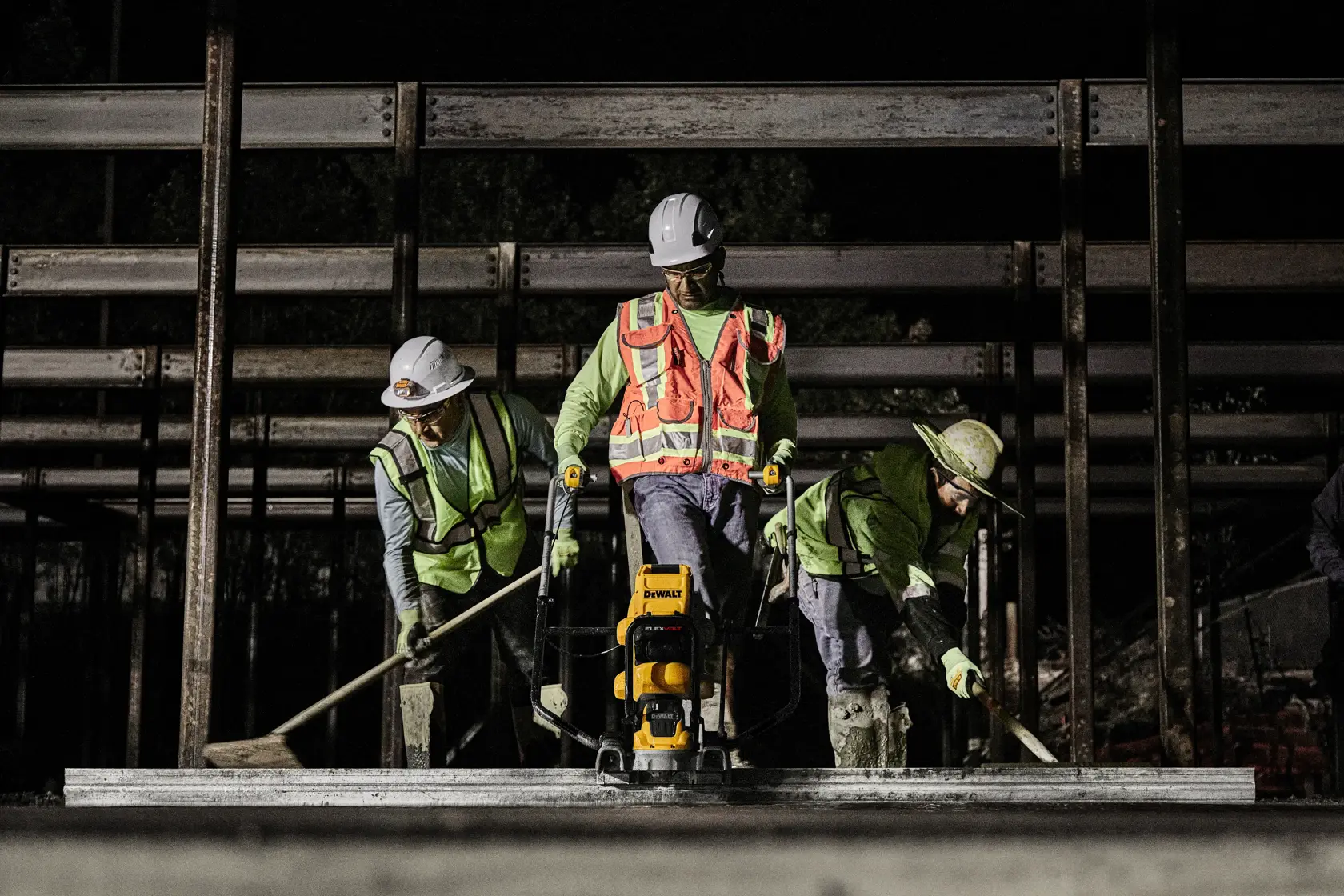 Three construction workers wearing safety vests and helmets are working at night on a building site. One worker uses a DEWALT power tool to smooth concrete, while the others assist with tools.