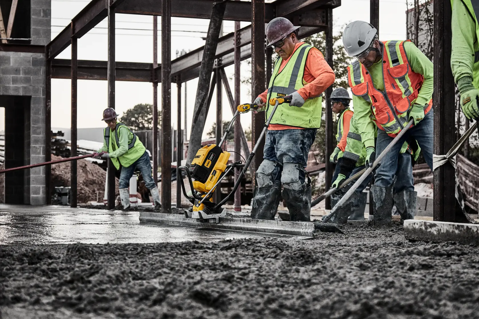 Several construction workers wearing safety vests and helmets are using a DEWALT concrete power screed to smooth out fresh concrete at a building site. The workers are operating the equipment and spreading the concrete in an unfinished structure.