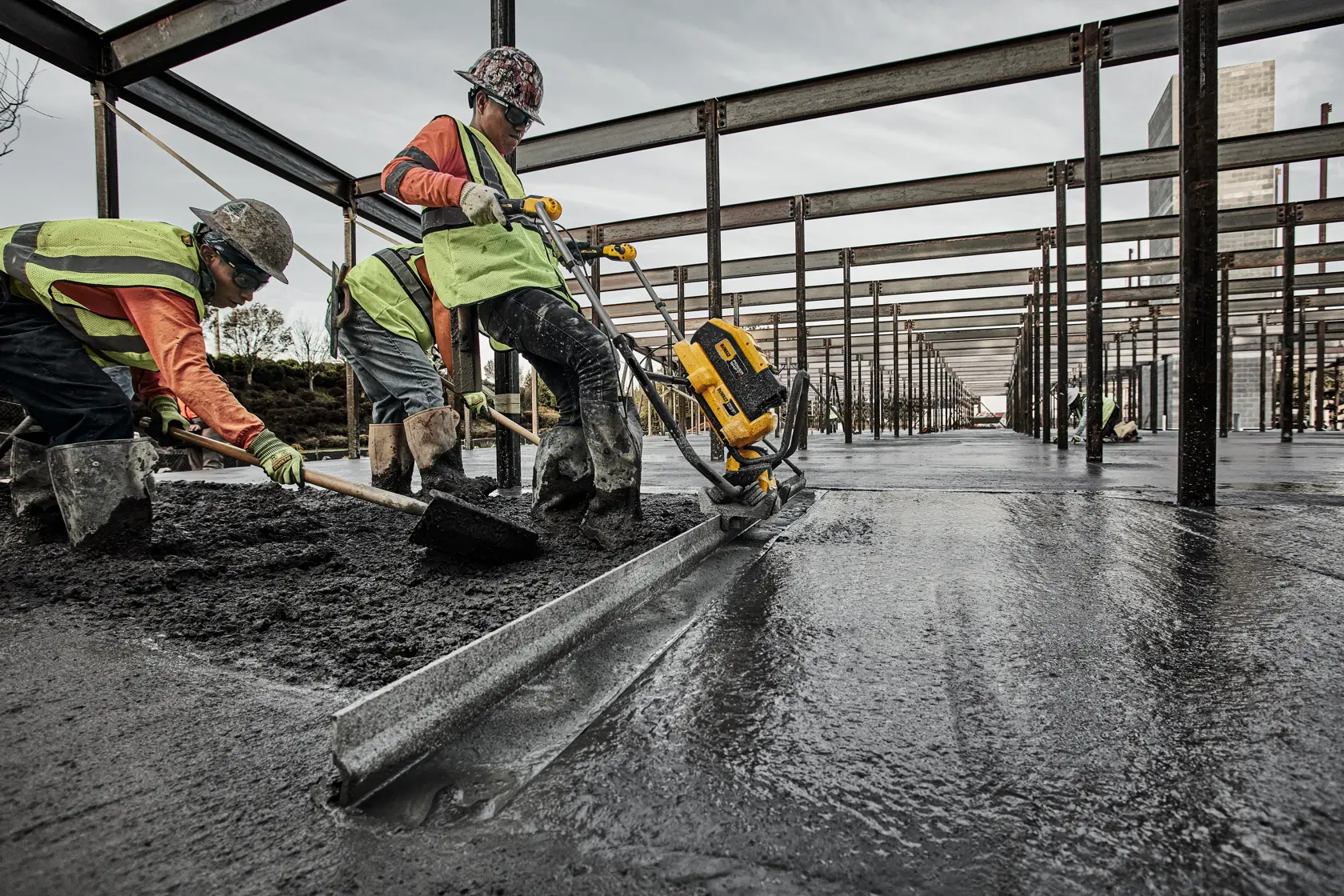 Workers wearing safety vests and helmets are leveling concrete at a construction site using a DEWALT power tool. The area is structured with steel beams, and the ground appears freshly poured and smoothed.