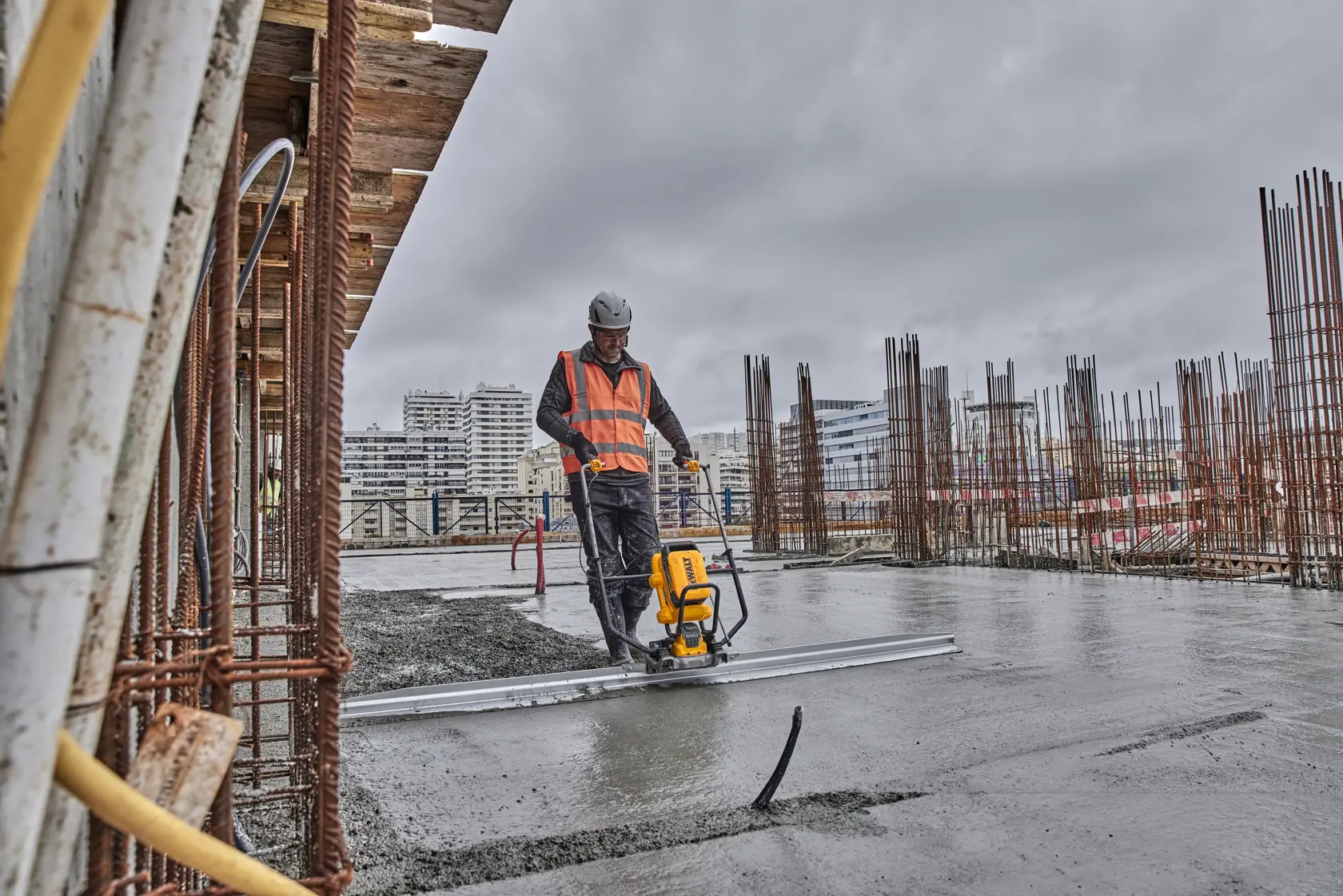 A person wearing a safety vest and helmet uses a DEWALT concrete screed machine to smooth a wet concrete surface at a construction site. Rebar rods and city buildings are visible in the background under a cloudy sky.