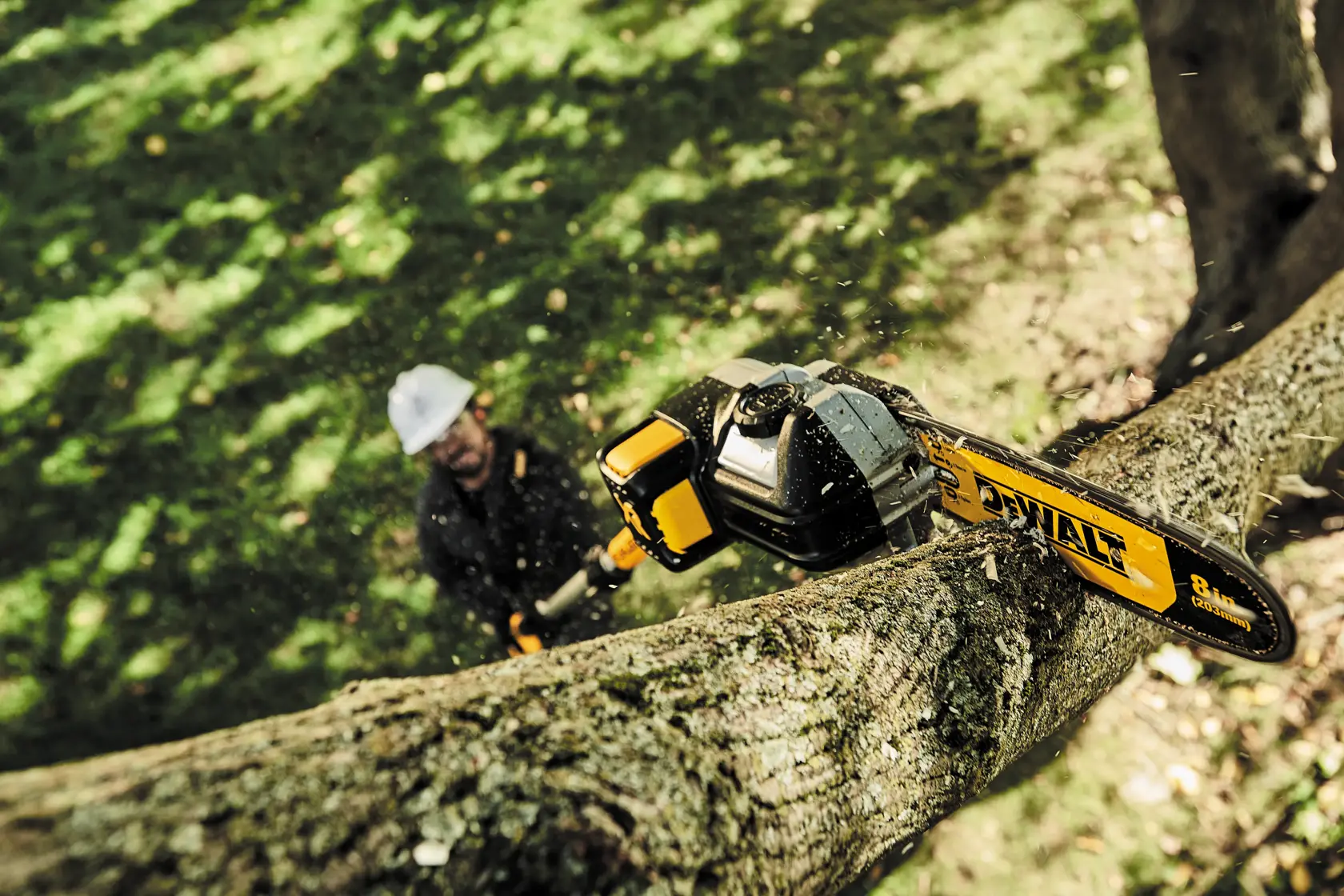 A DEWALT pole saw cutting through a tree branch in an outdoor setting. There is a person in the background operating the tool, but their face is blurred.