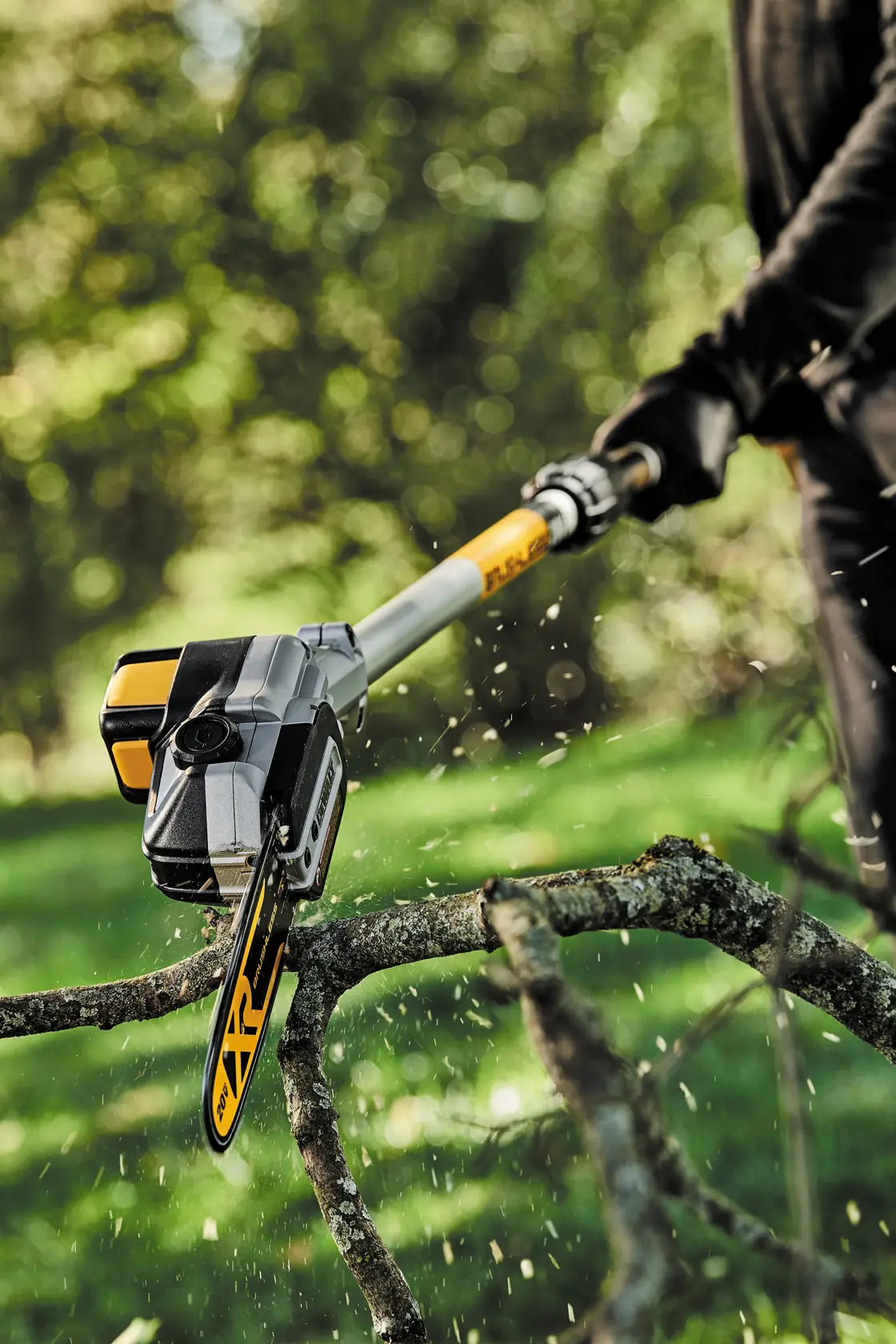 A DEWALT DCPS620M1 pole saw being used to cut a tree branch outdoors, with sawdust particles visible in the air.