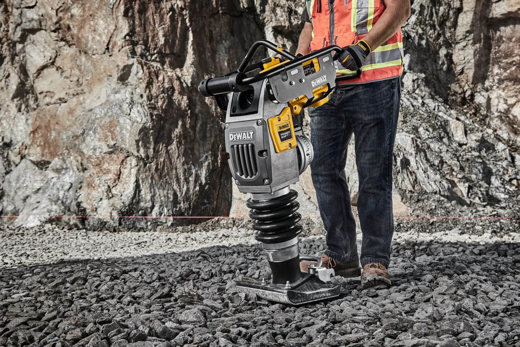 A person using a DEWALT compactor tool on a rocky surface at a construction site, wearing safety gear including a high-visibility vest, gloves, and work boots.