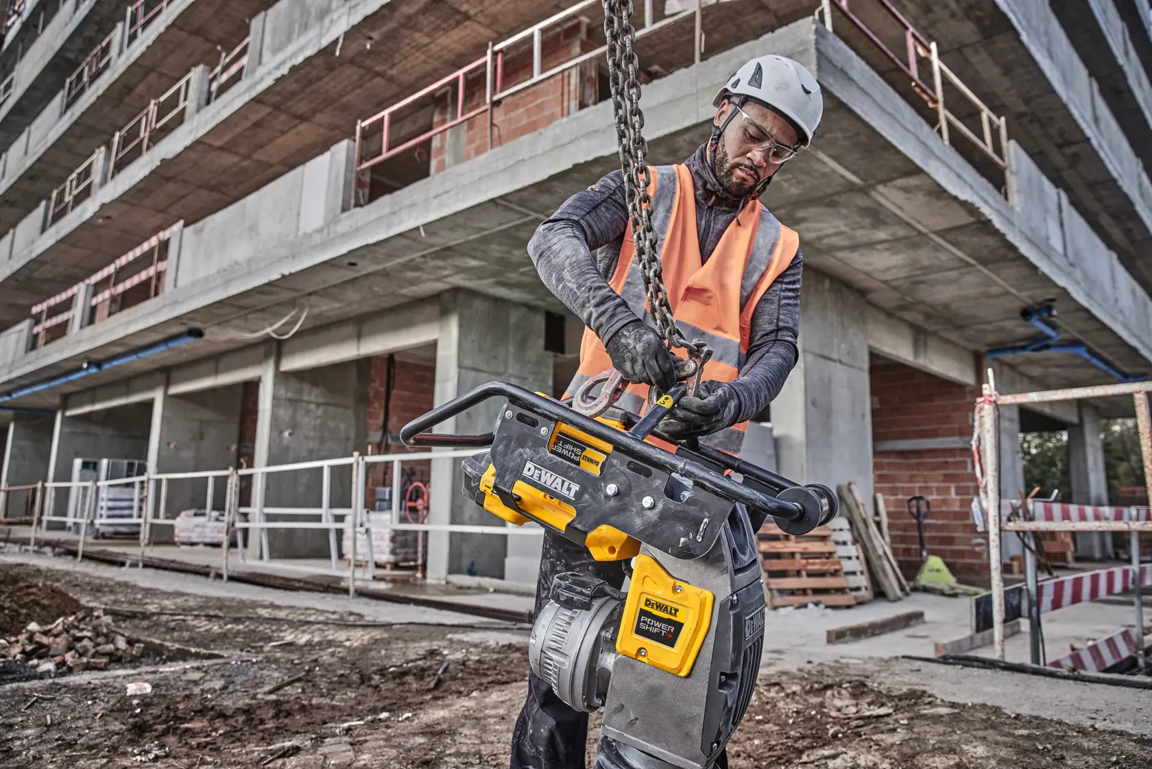 A worker in safety gear is using a DEWALT power tool at a construction site in front of a multi-story concrete building. The worker's face is blurred for privacy.
