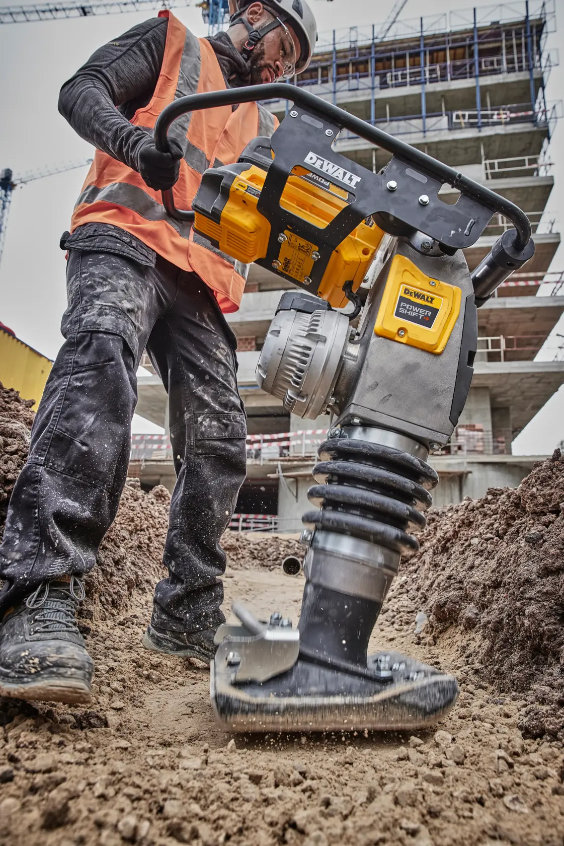 A construction worker wearing a safety vest and helmet operates a DEWALT DCPS660AG2 compactor on a dirt pathway at a building site. The DEWALT device is shown in close detail, actively compacting soil.