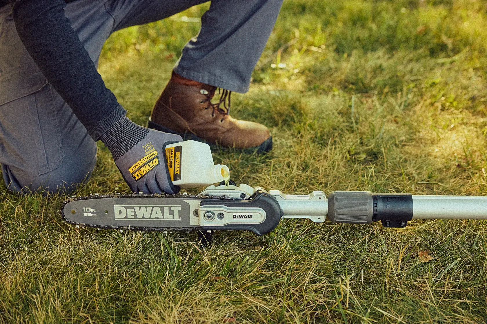 A person wearing gloves and work boots is pouring oil into a DEWALT pole saw resting on grass.
