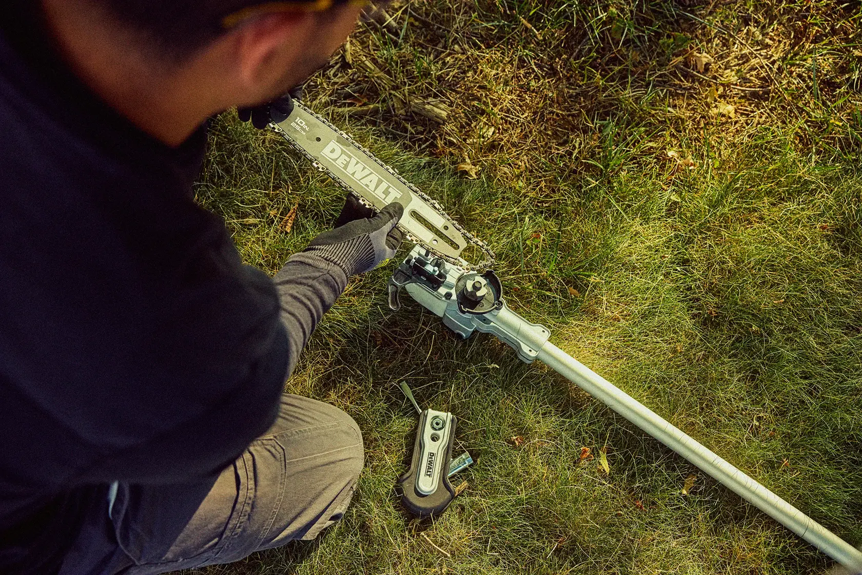 A person wearing gloves is assembling or adjusting a DEWALT pole saw on grass. The tool has a long pole and a saw attachment at the end, with power tool parts visible on the ground.