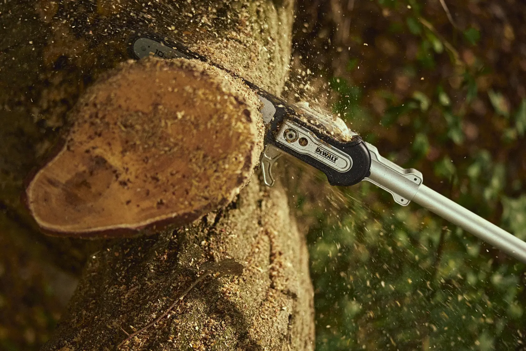 A DEWALT pole saw cutting through a thick tree branch with wood chips flying, outdoors in a forest setting.