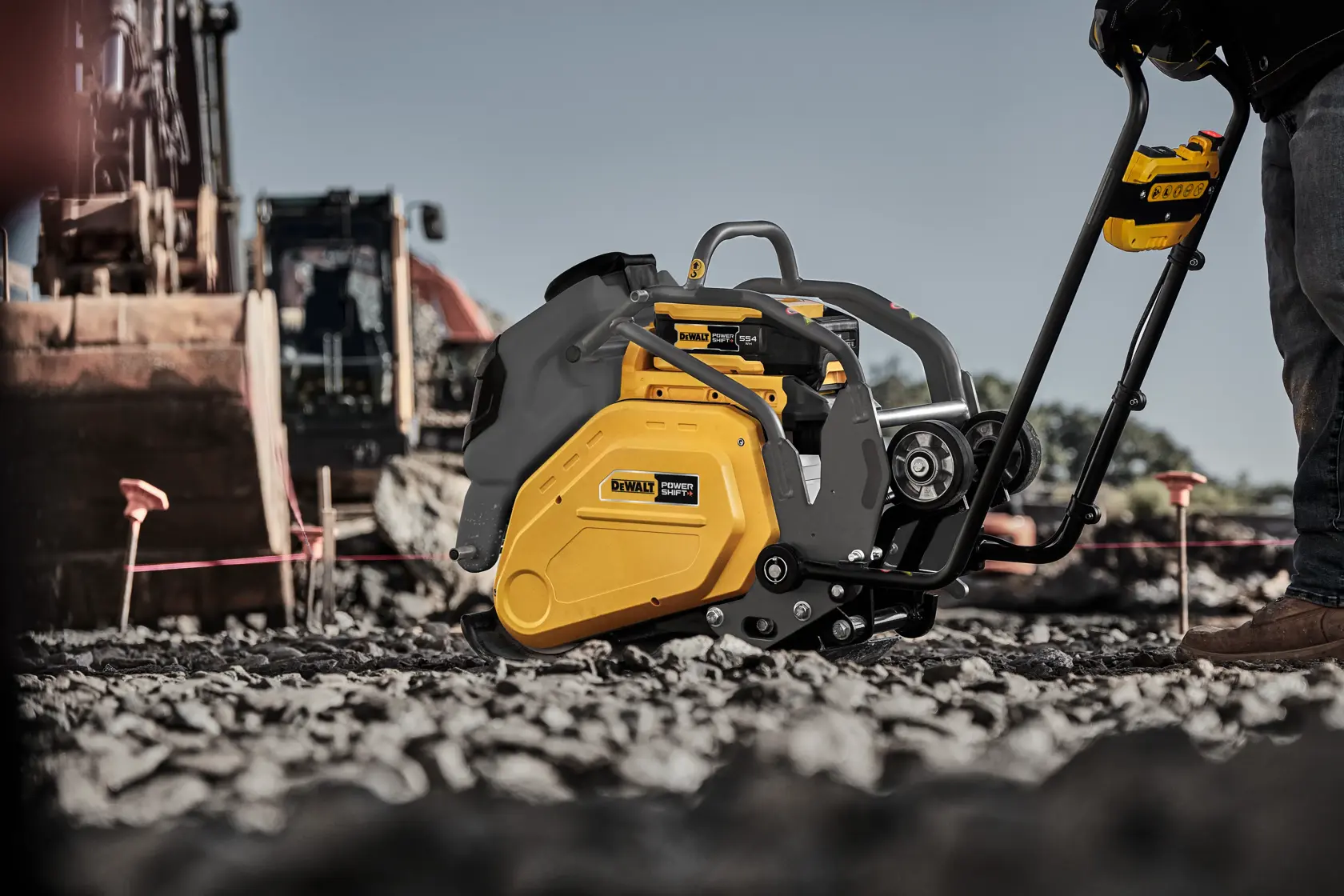 A DEWALT compact plate compactor is being used on a rocky outdoor construction site. The yellow and black compactor is positioned on loose gravel with part of a construction vehicle visible in the background.