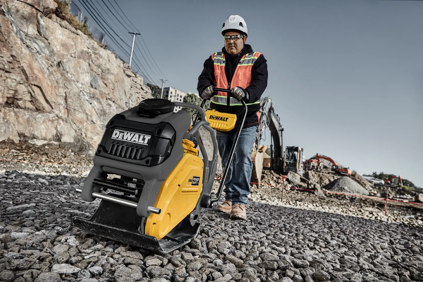 A construction worker operating a DEWALT plate compactor on a gravel surface at a construction site. The worker is wearing safety gear, including a helmet and orange safety vest. There is construction equipment and rocky terrain in the background.