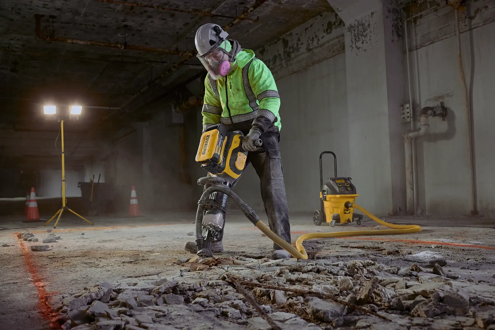 A construction worker in safety gear, including a helmet, gloves, high-visibility jacket, and respiratory mask, operates a yellow industrial jackhammer to break up a concrete floor in a dimly lit underground area. Debris and rubble surround the worker, and orange safety cones and floodlights are visible in the background.