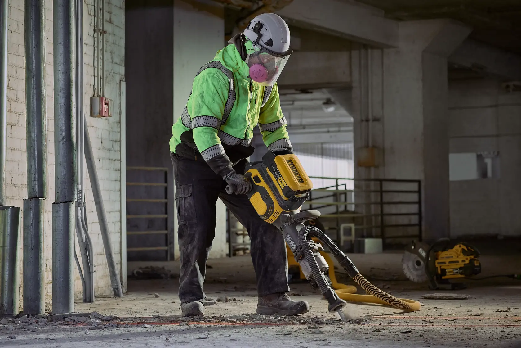 A person wearing safety gear, including a helmet, mask, and reflective jacket, operates a yellow and black industrial jackhammer inside a construction site with concrete floors and exposed pipes.