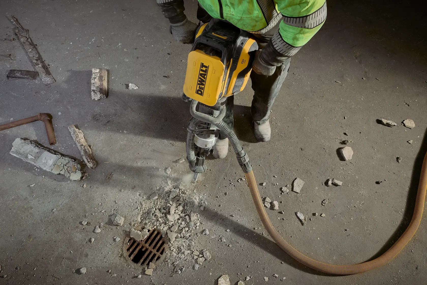 A construction worker using a yellow DEWALT jackhammer to break up concrete near a floor drain, with rubble and debris scattered around.