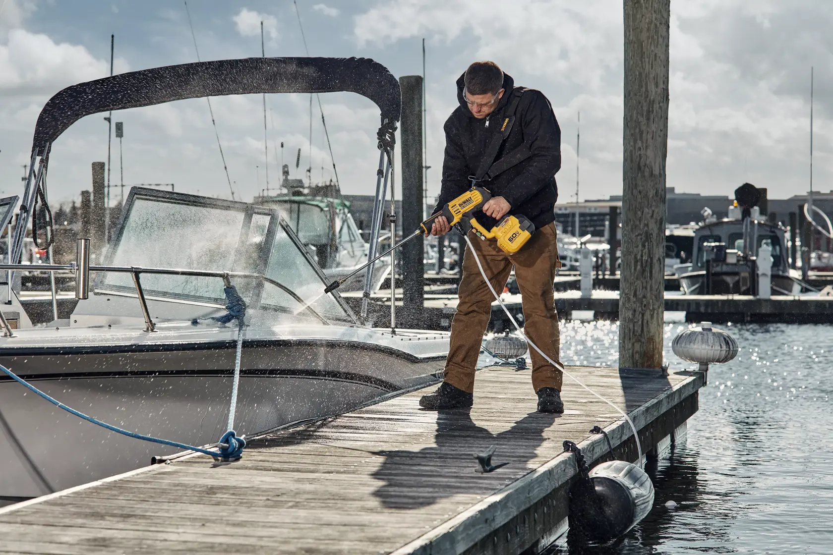 A person is using a DEWALT cordless power washer (DCPW1000X1) to clean a boat at a dock. The person is standing on a wooden dock, spraying water onto the side of the boat, with marina and boats in the background.
