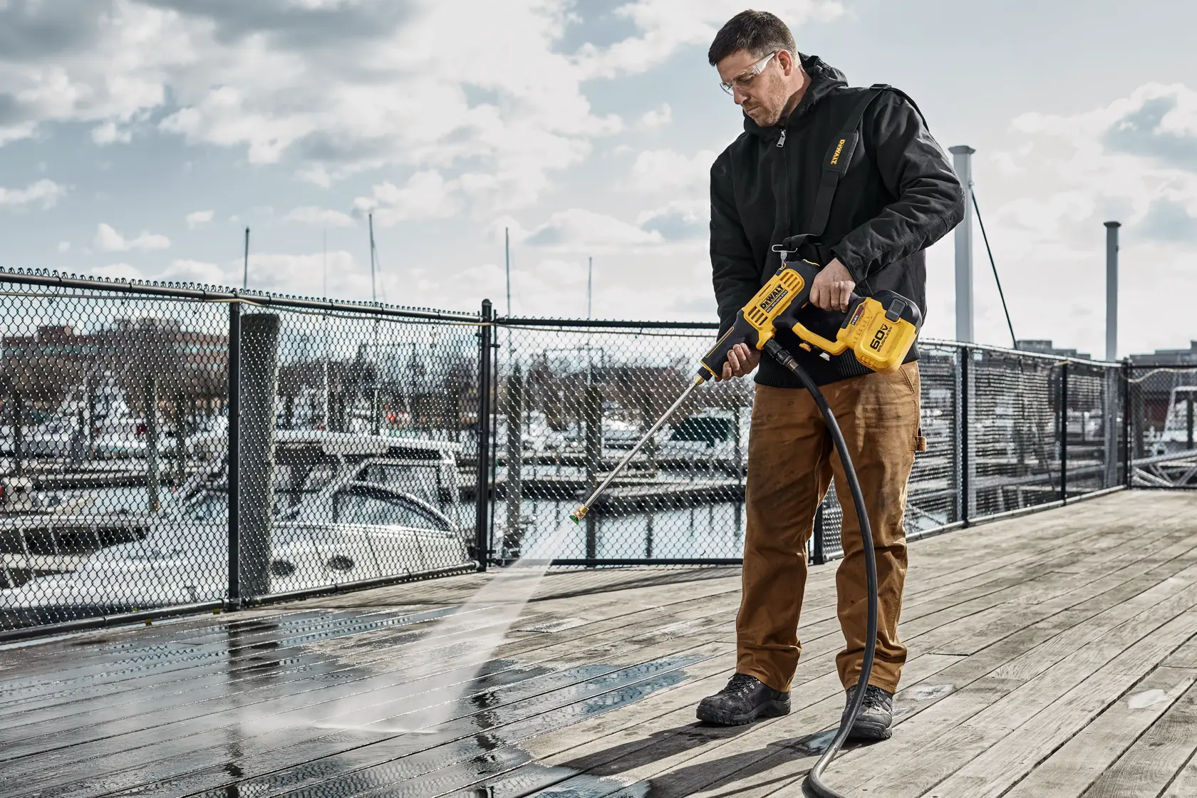 A person is using a DEWALT cordless power cleaner to wash a wooden dock next to a marina with boats in the background. The power cleaner is yellow and black, and water is spraying from the nozzle onto the dock.
