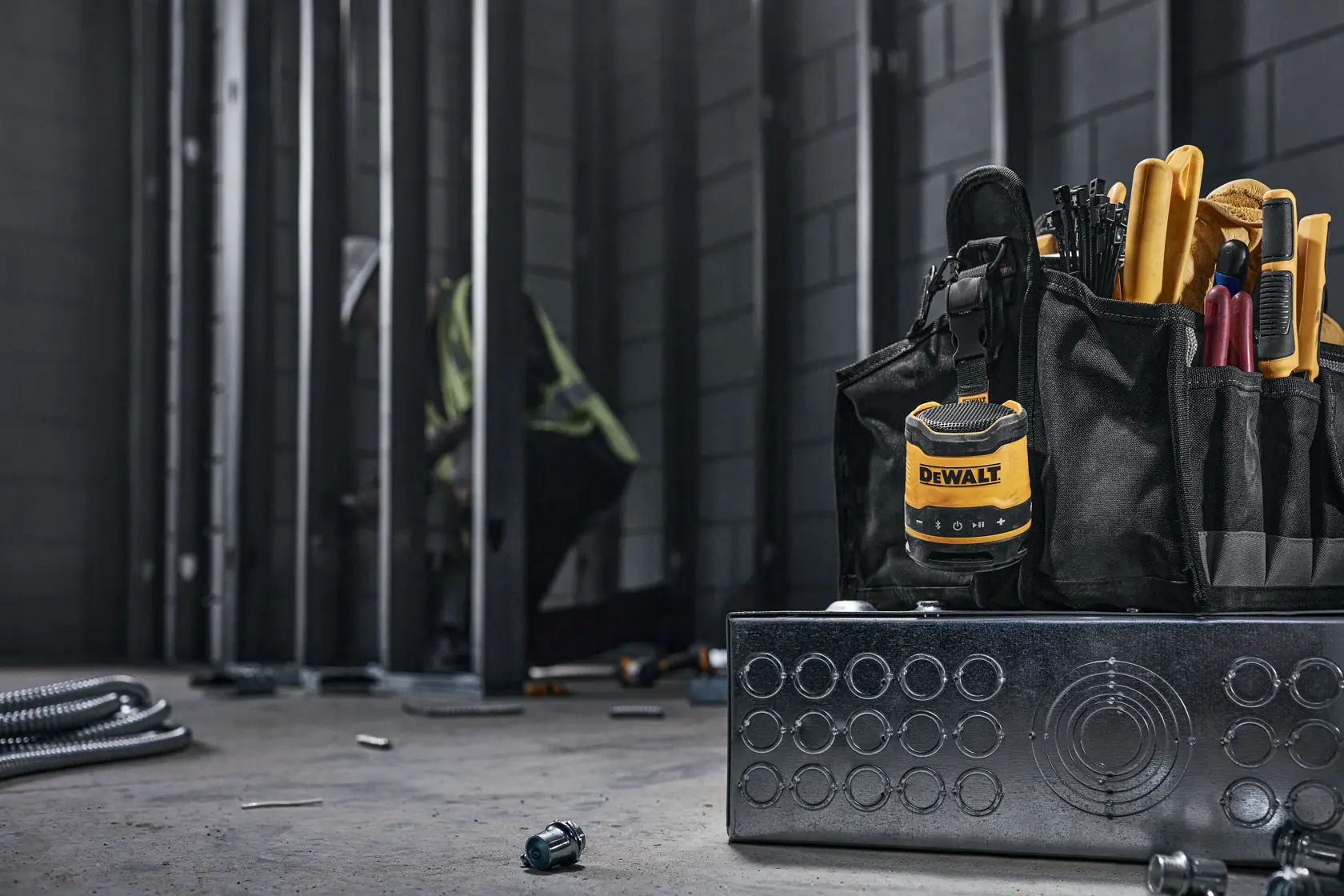 A DEWALT portable Bluetooth speaker (model DCR008) attached to a black tool bag filled with various hand tools, set in an industrial construction environment with metal framing and scattered hardware on the floor.