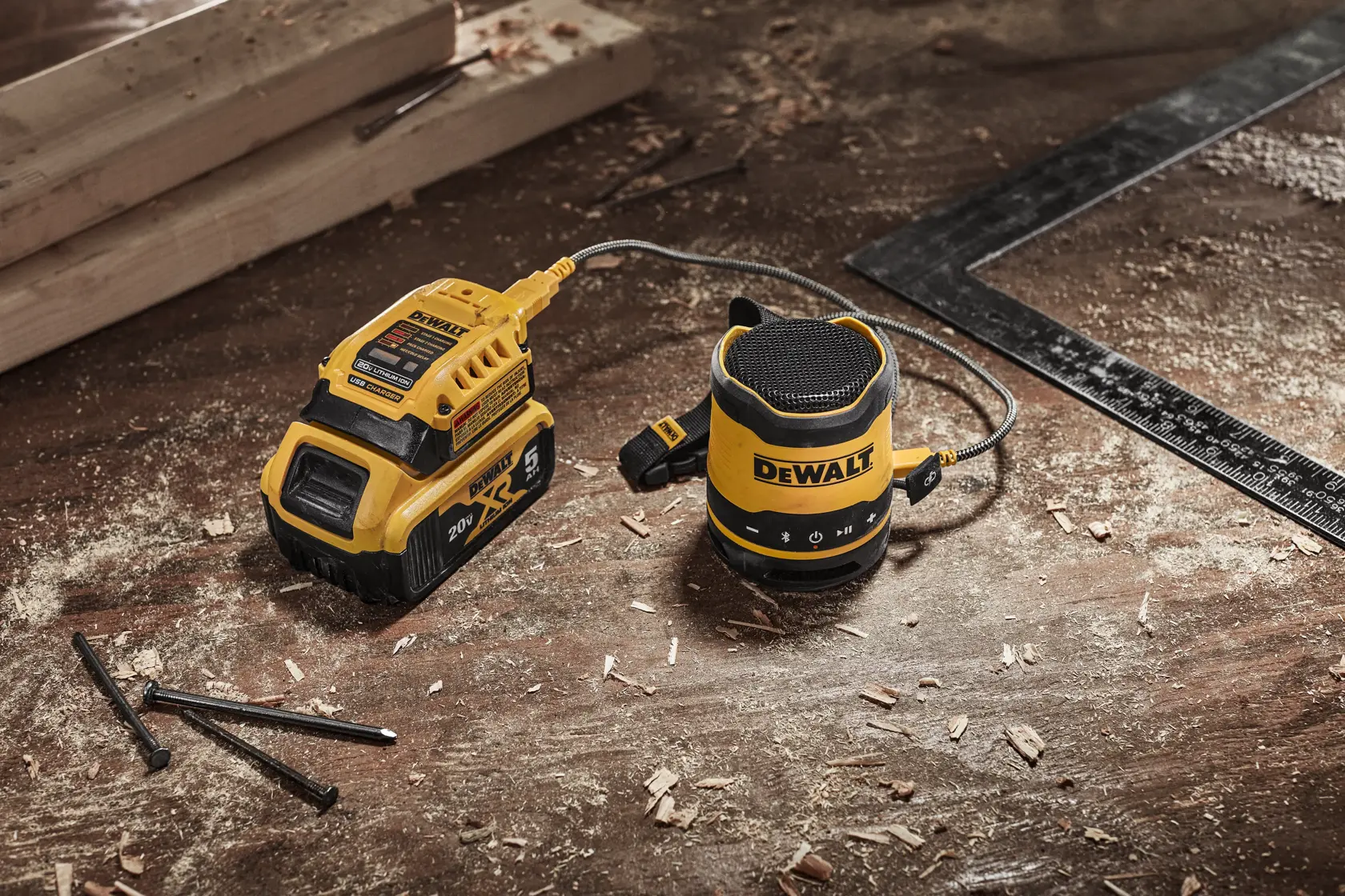 A DEWALT portable Bluetooth speaker connected to a DEWALT 20V battery, placed on a workbench with wood shavings, nails, and a carpenter's square.