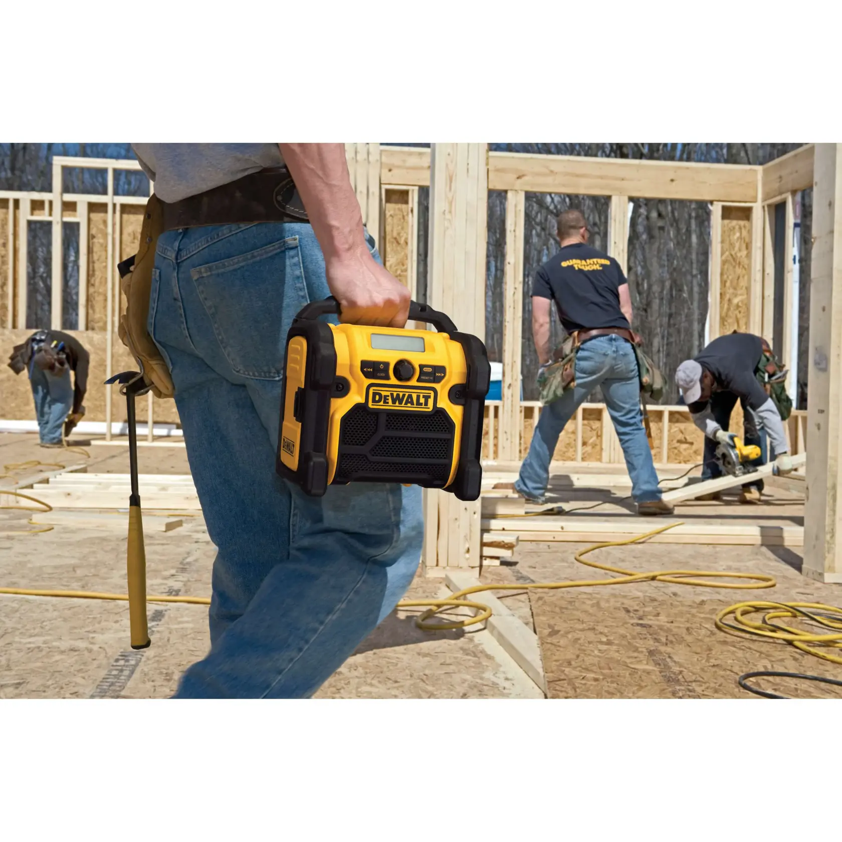 A person is holding a yellow DEWALT portable radio, model DCR018, on a construction site with several workers building a wooden framework.