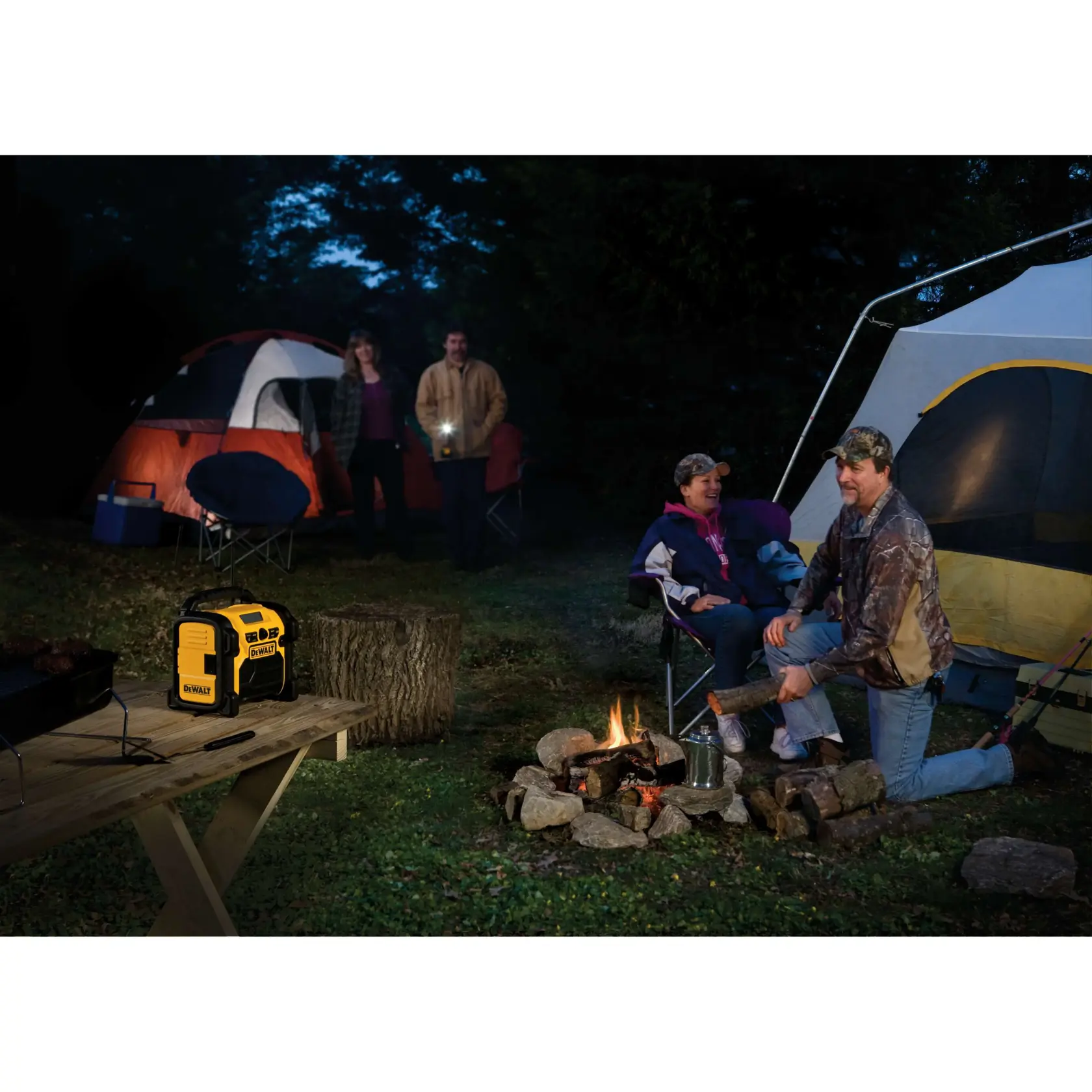 A DEWALT portable radio sits on a wooden picnic table at a nighttime campsite. Two tents are set up, with two people standing near one tent and two people sitting by a campfire near the other tent. The scene includes camping chairs, firewood, and outdoor cooking gear.