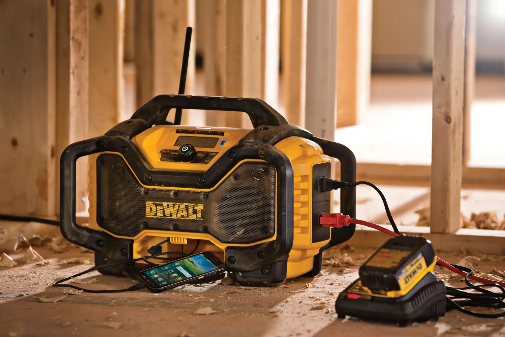A DEWALT jobsite radio with built-in charger is placed on a construction site floor. A smartphone is connected and sits underneath in a compartment, and a DEWALT battery charger is plugged in beside it. The radio features rugged protective bumpers.