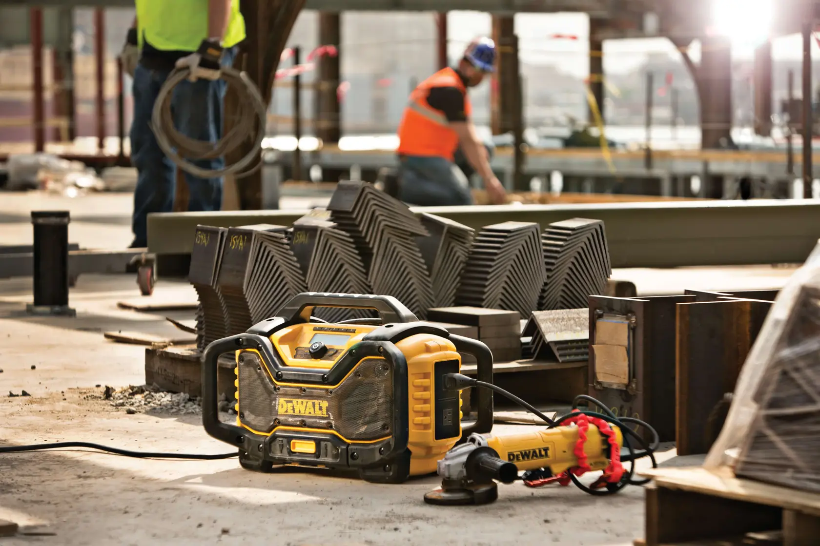 A DEWALT DCR025 jobsite radio placed on the ground at a construction site, surrounded by steel materials and tools, with workers and industrial equipment visible in the background.