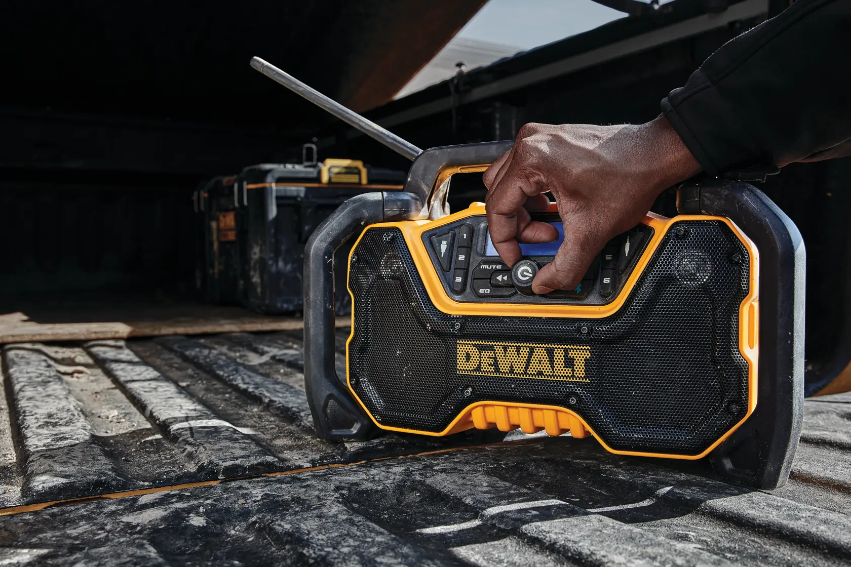 A close-up of a person's hand adjusting the settings on a DEWALT portable radio placed on the bed of a truck. The radio has a rugged design and is black and yellow, with other tools visible in the background.