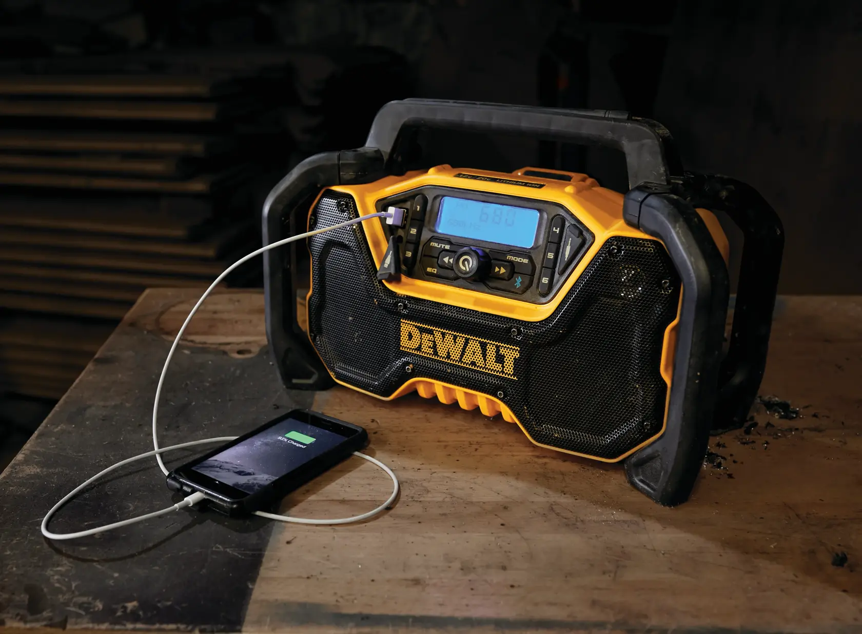 A DEWALT portable radio with a digital display is placed on a wooden workbench. A smartphone is charging via a cable connected to the radio. The scene appears to be in a workshop with stacked wood in the background.
