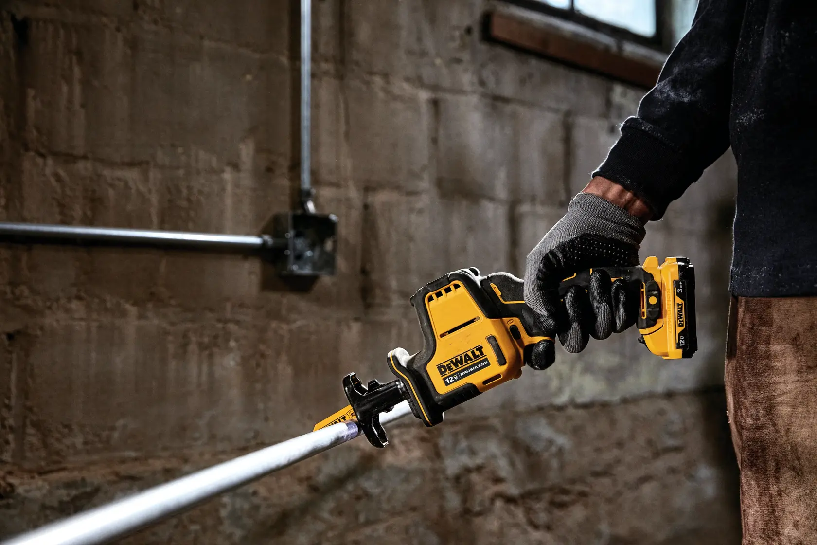 A person wearing gloves is holding and using a DEWALT DCS312B cordless reciprocating saw to cut a metal pipe in an industrial or workshop setting.