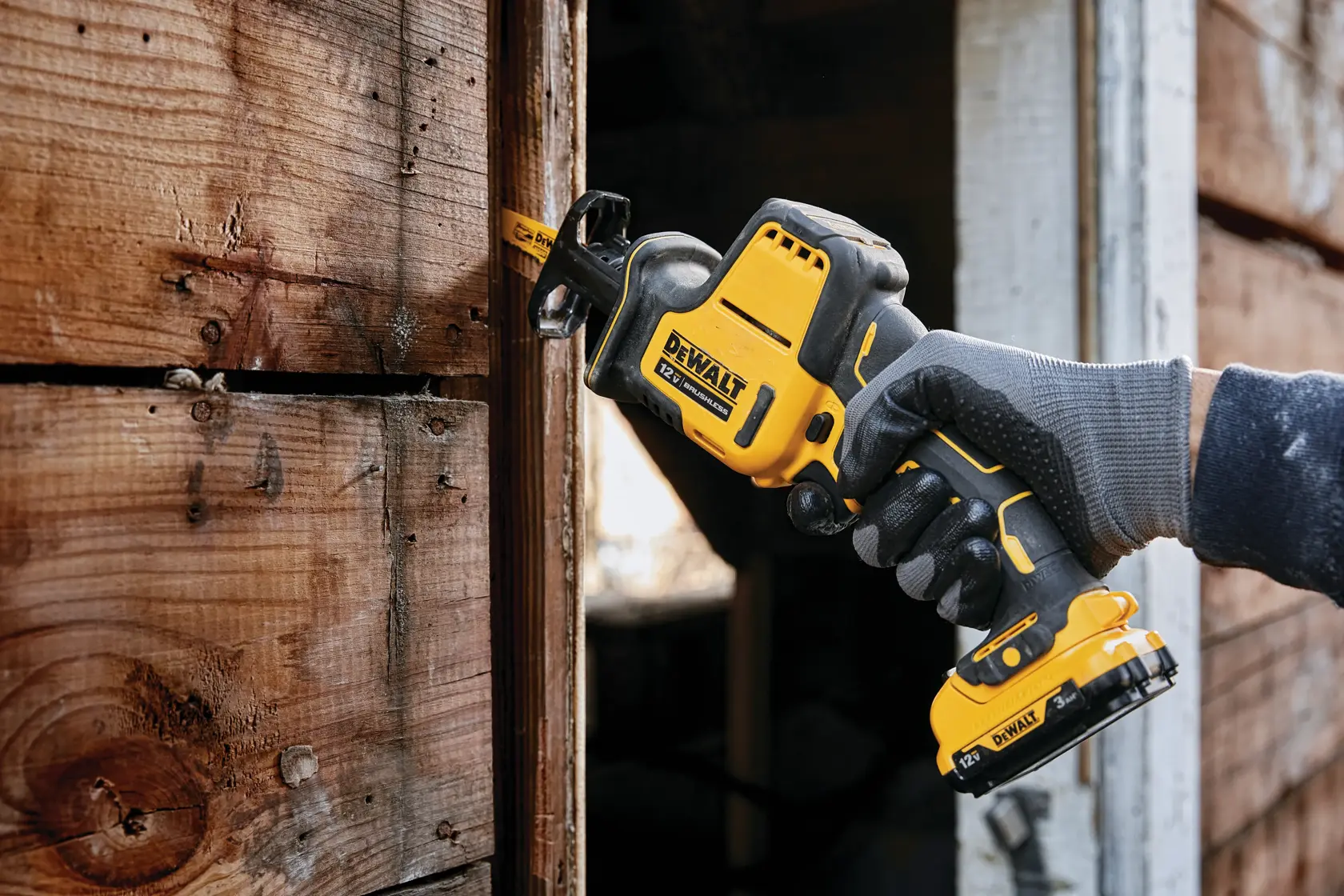 A person wearing gloves uses a DEWALT cordless reciprocating saw (model DCS312B) to cut through a wooden plank near a doorway.
