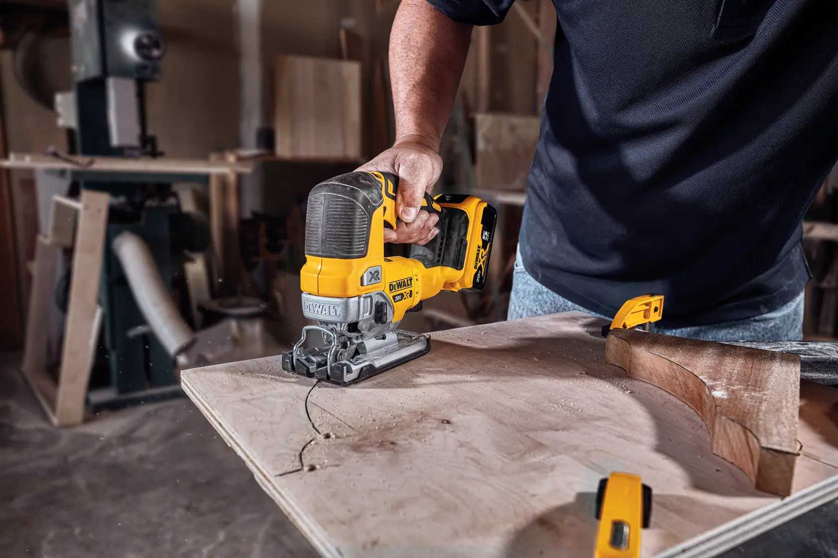 A person using a DEWALT cordless jigsaw to cut a curved line in a piece of wood in a workshop.