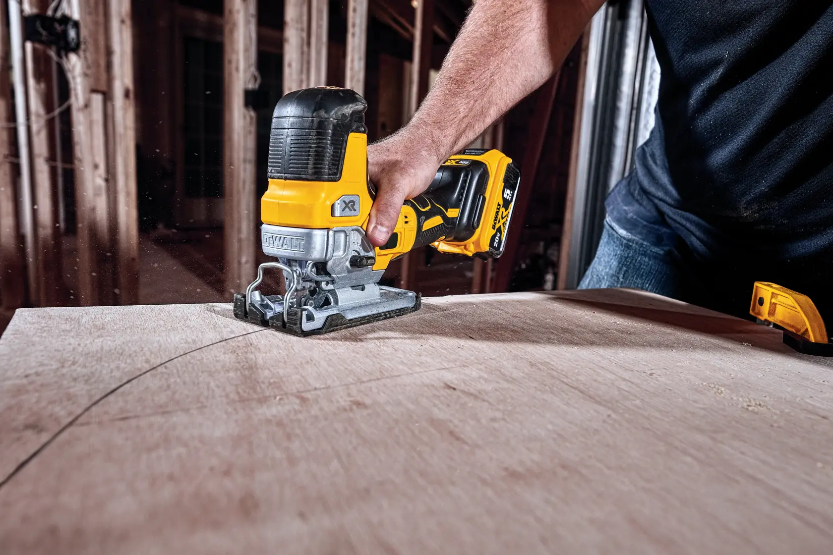 A person using a DEWALT cordless jigsaw to cut a curved line in a piece of wood inside a construction site.