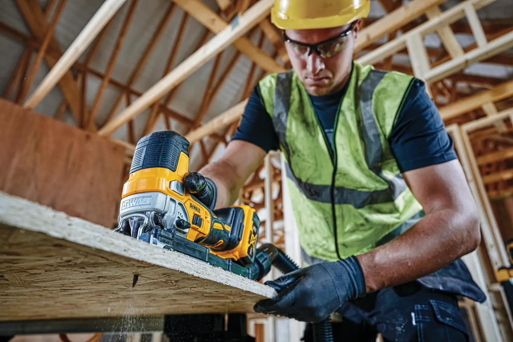 A construction worker wearing a safety vest and gloves uses a DEWALT DCS335B cordless jigsaw to cut a piece of wood at a building site with exposed beams.