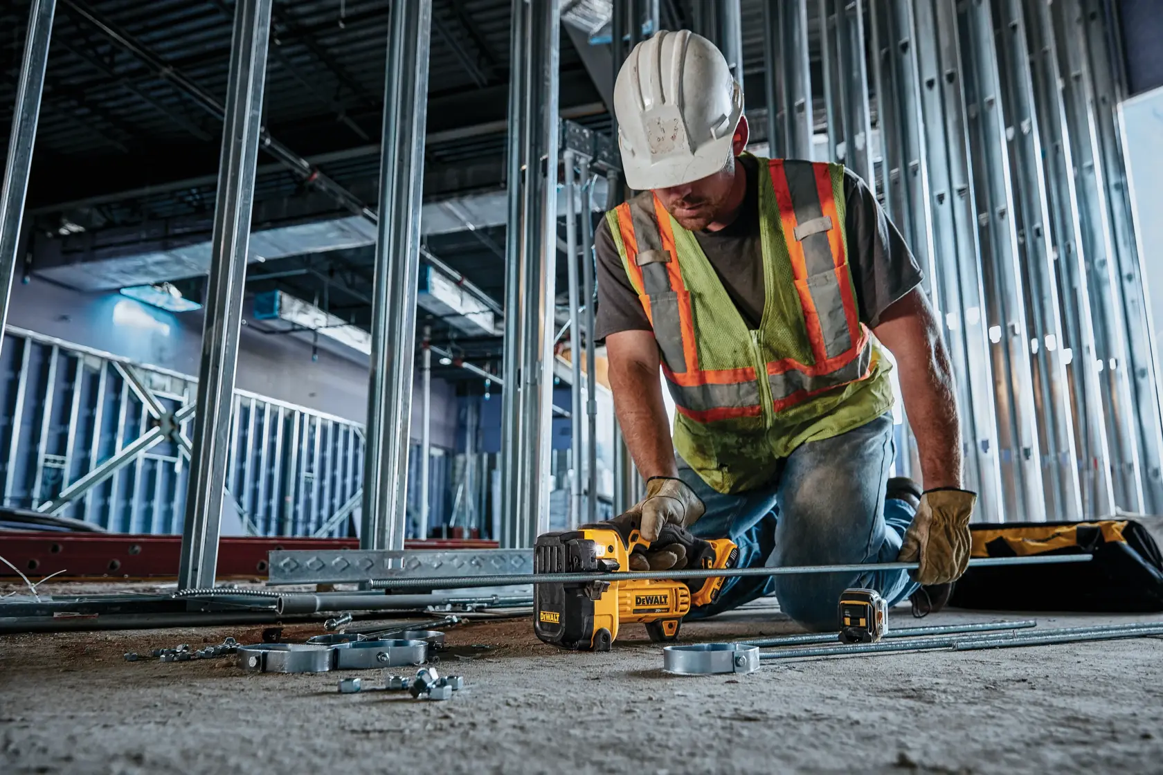 A construction worker wearing a safety vest, gloves, and a hard hat uses a DEWALT DCS350B tool to cut metal rods at a building site, surrounded by metal framing and construction supplies.