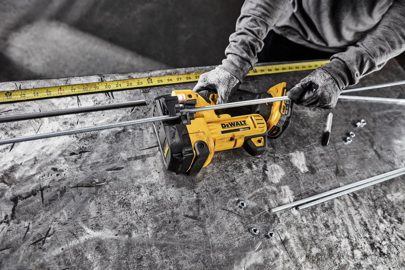 A person wearing gloves uses a yellow DEWALT cordless threaded rod cutter to cut metal rods on a workbench. Several rods, nuts, and a measuring tape are visible on the table.