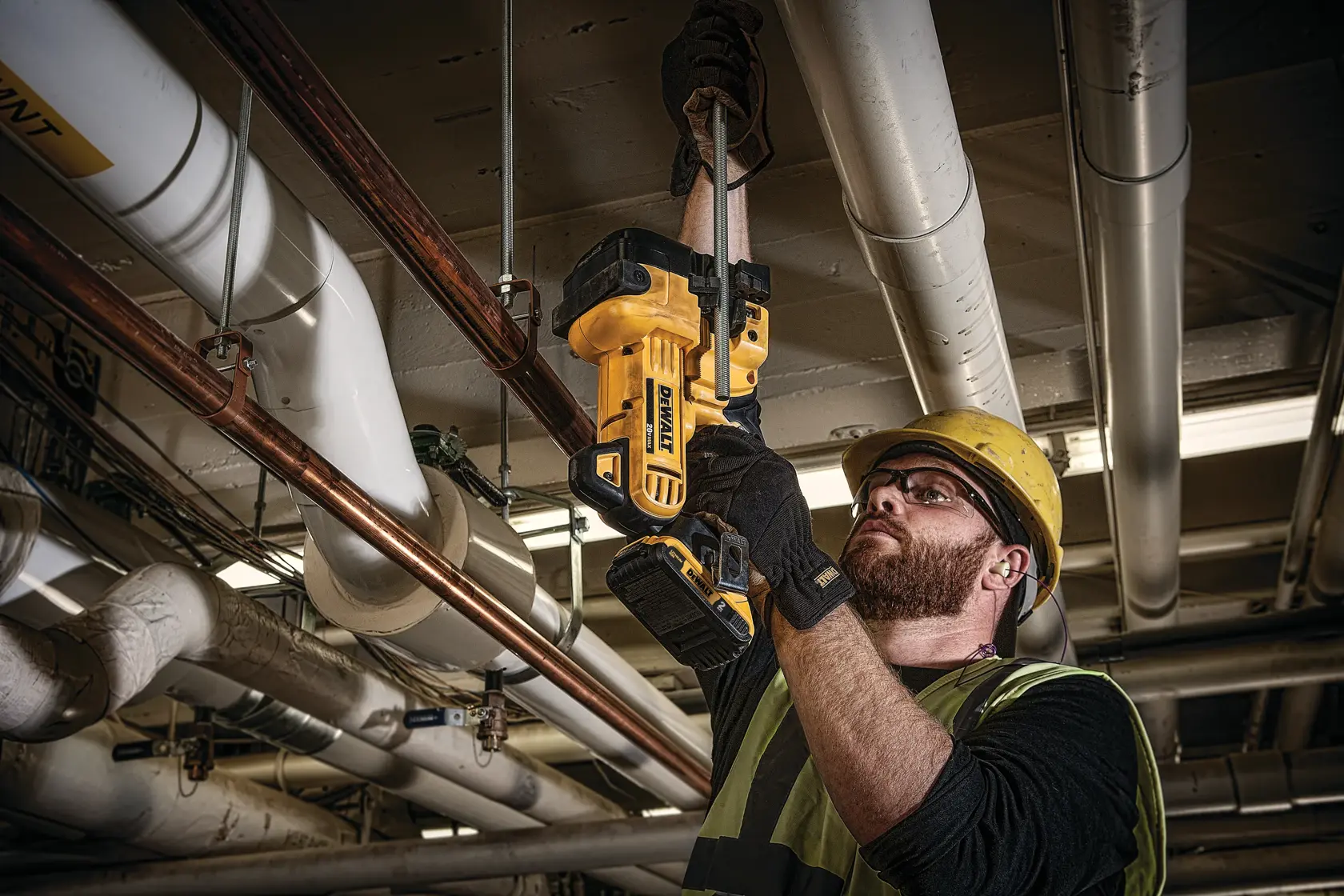 A worker wearing safety gloves and a helmet uses a DEWALT DCS350D1 cordless tool to cut through a metal rod in an industrial setting beneath pipes.