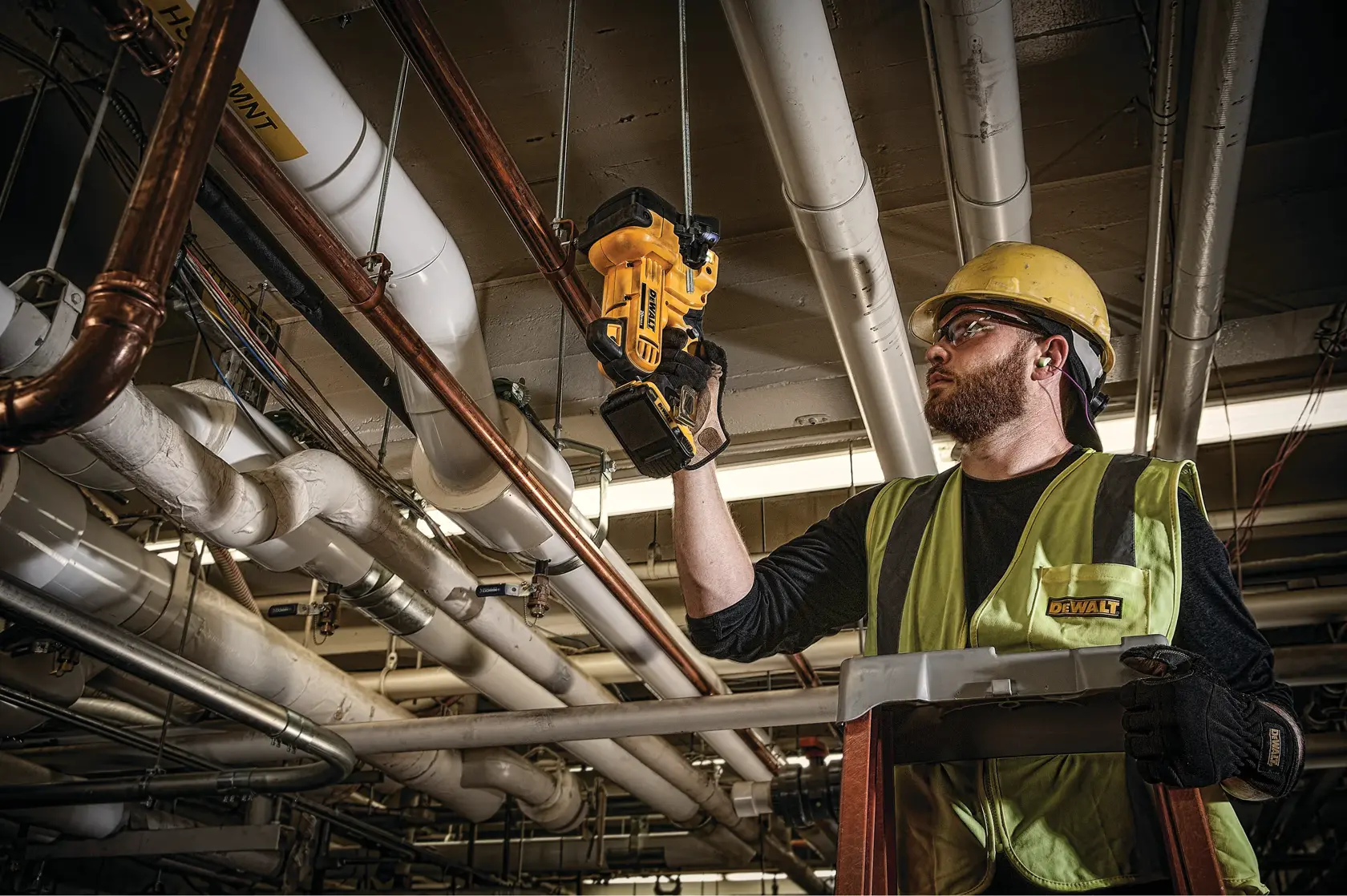 A person wearing a yellow hard hat and safety vest uses a DEWALT cordless power tool to work on ceiling pipes and conduits. The person is standing on a ladder in an industrial setting.