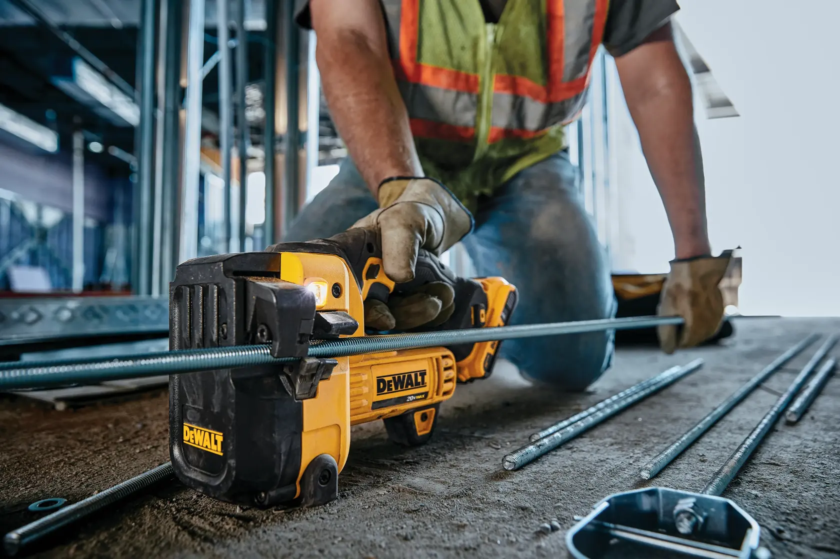A worker using a DEWALT cordless threaded rod cutter tool to cut a metal rod at a construction site. The worker is wearing gloves and a safety vest.