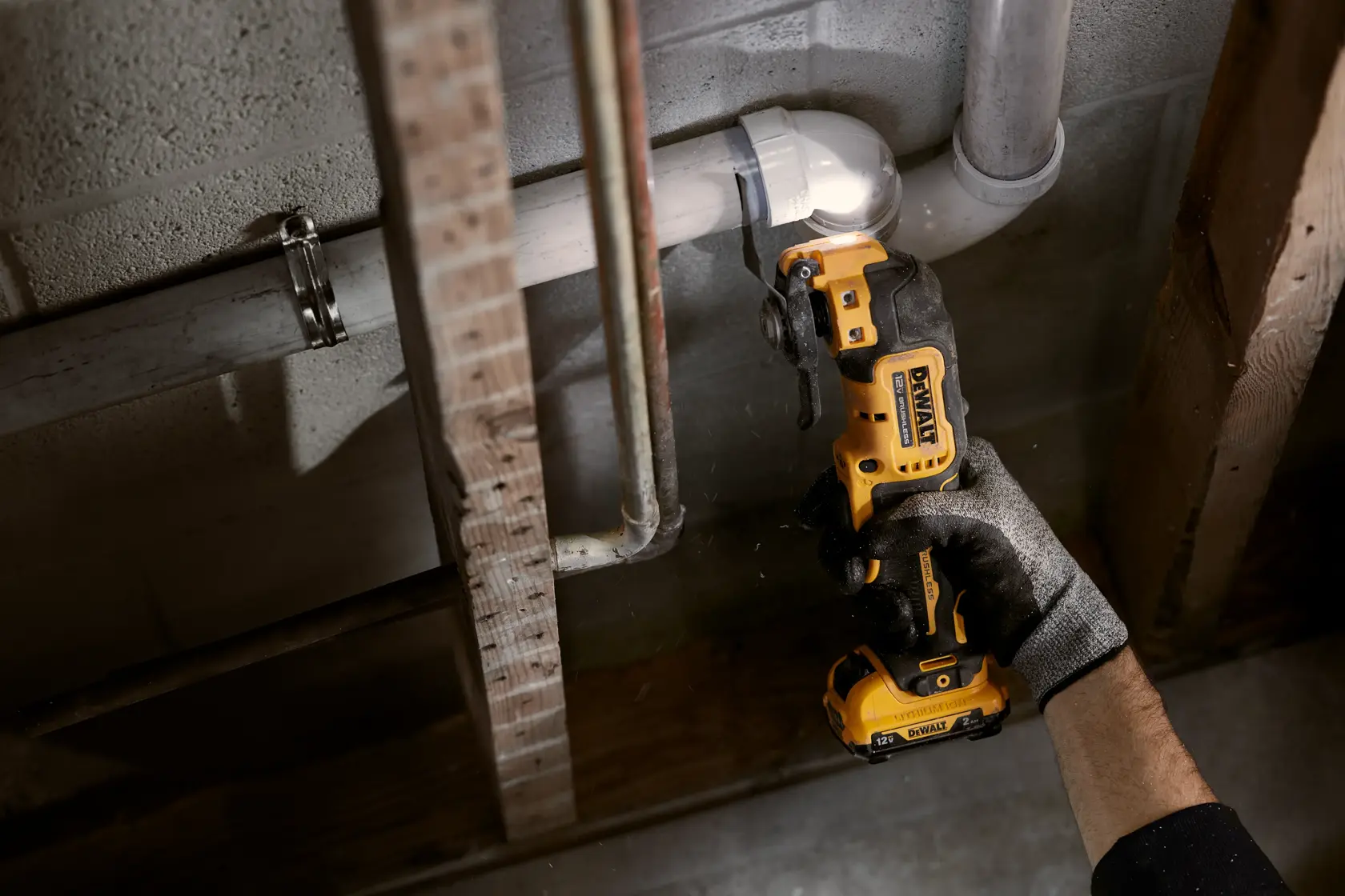 A person wearing a glove is using a DEWALT cordless oscillating multi-tool to cut a white PVC pipe near a wall with exposed plumbing.