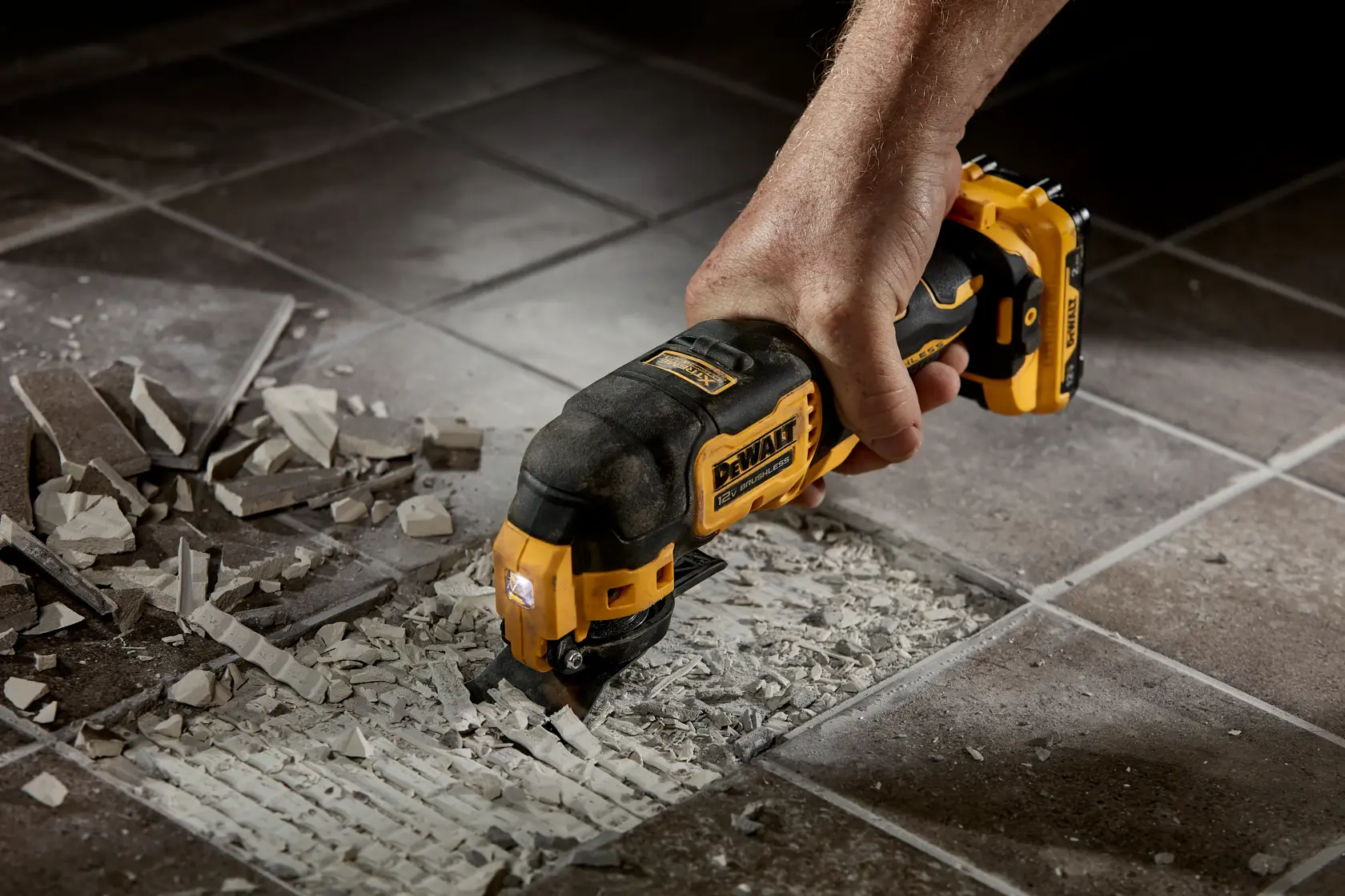 A hand holding a DEWALT cordless oscillating multi-tool (model DCS353B) removing and cutting out grout between tiles on a tiled floor, with broken grout and tile debris visible.