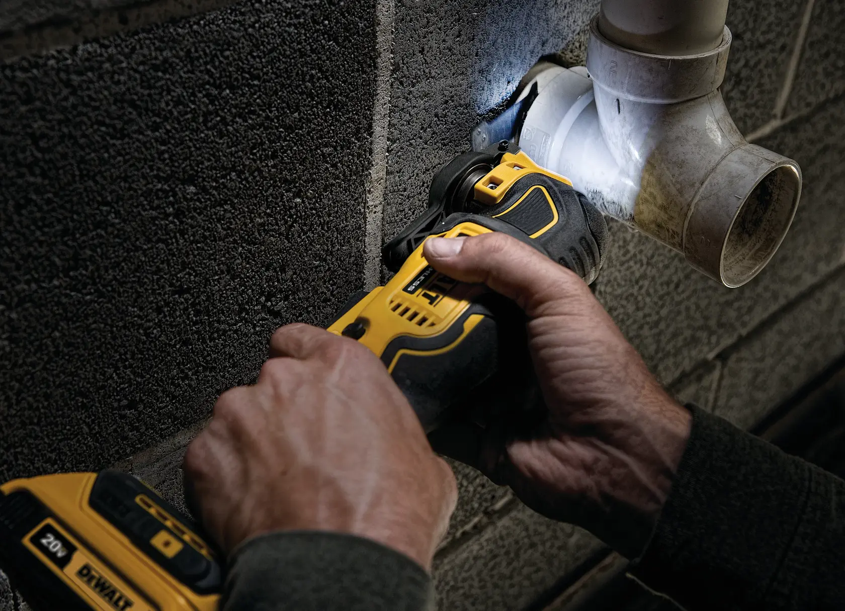 A person uses a DEWALT cordless oscillating tool to cut or trim material near a plastic pipe mounted on a concrete wall.